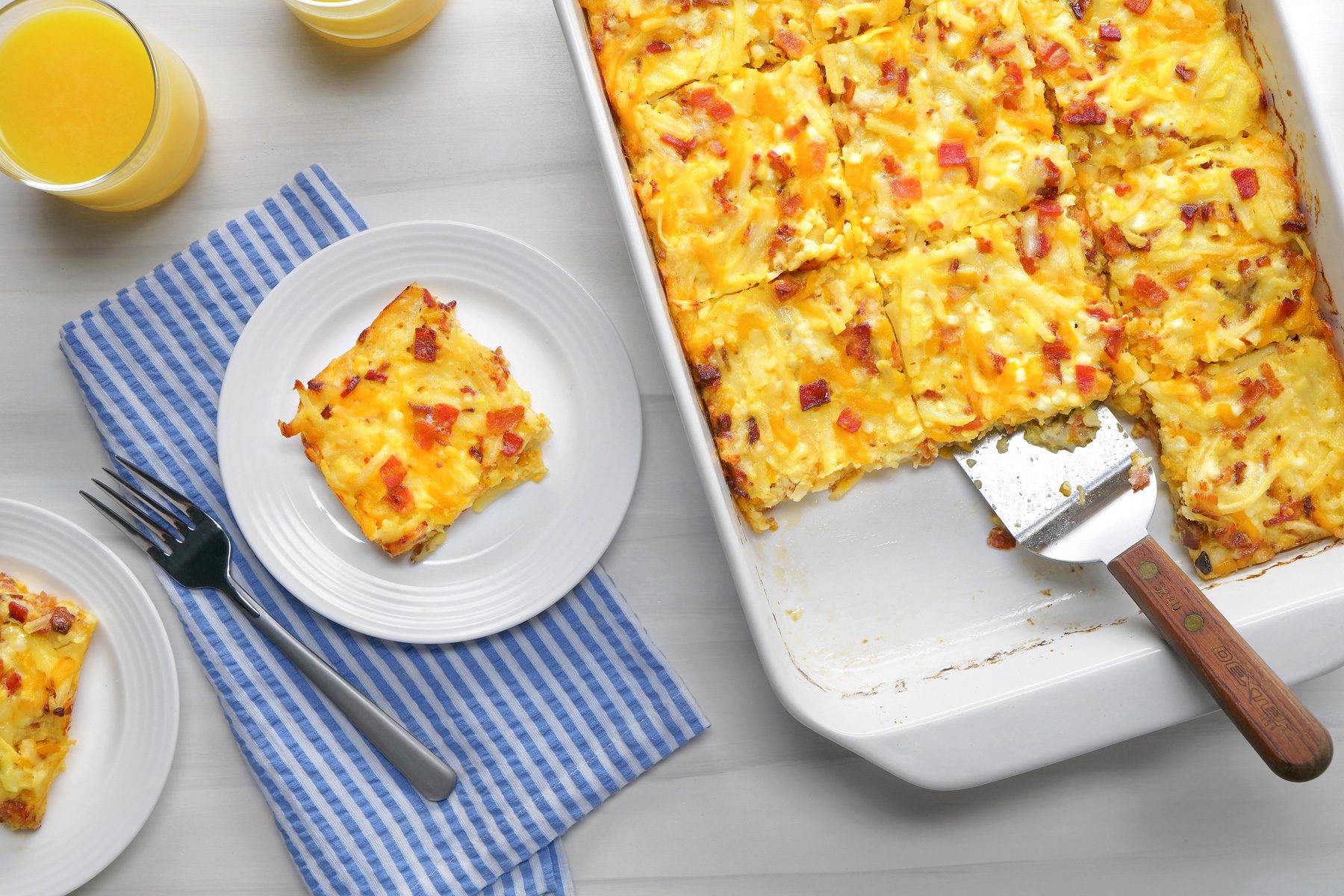 overhead shot; white background; Amish Breakfast Casserole in small white plate; Freshly cut squares are arranged on white plates beside the main dish; The casserole is nestled in a white baking dish, with a spatula resting on the edge; Two glasses of orange juice and a blue and white striped napkin