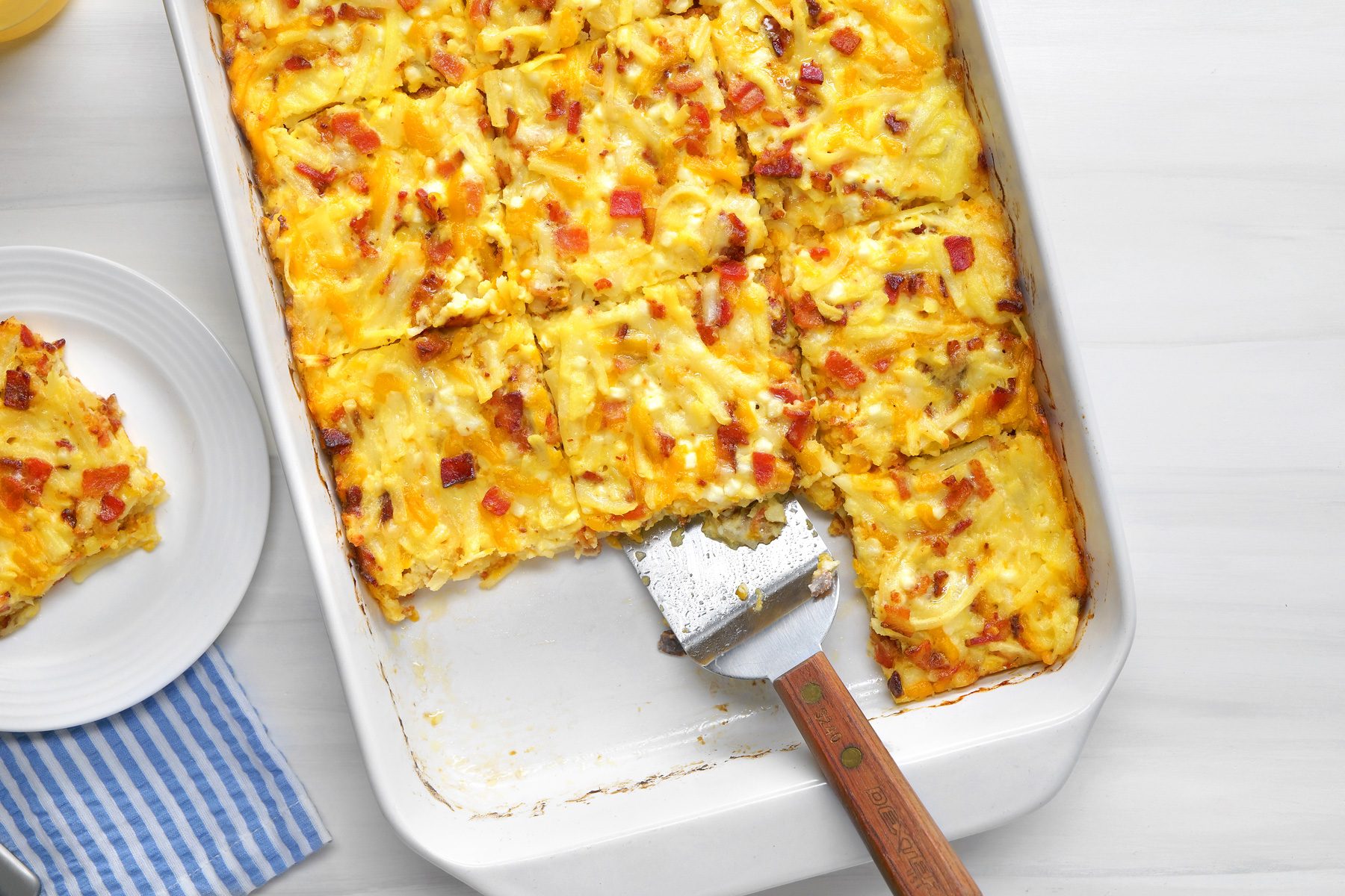 overhead shot; white background; Amish Breakfast Casserole in small white plate; Freshly cut squares are arranged on white plates beside the main dish; The casserole is nestled in a white baking dish, with a spatula resting on the edge; Two glasses of orange juice and a blue and white striped napkin