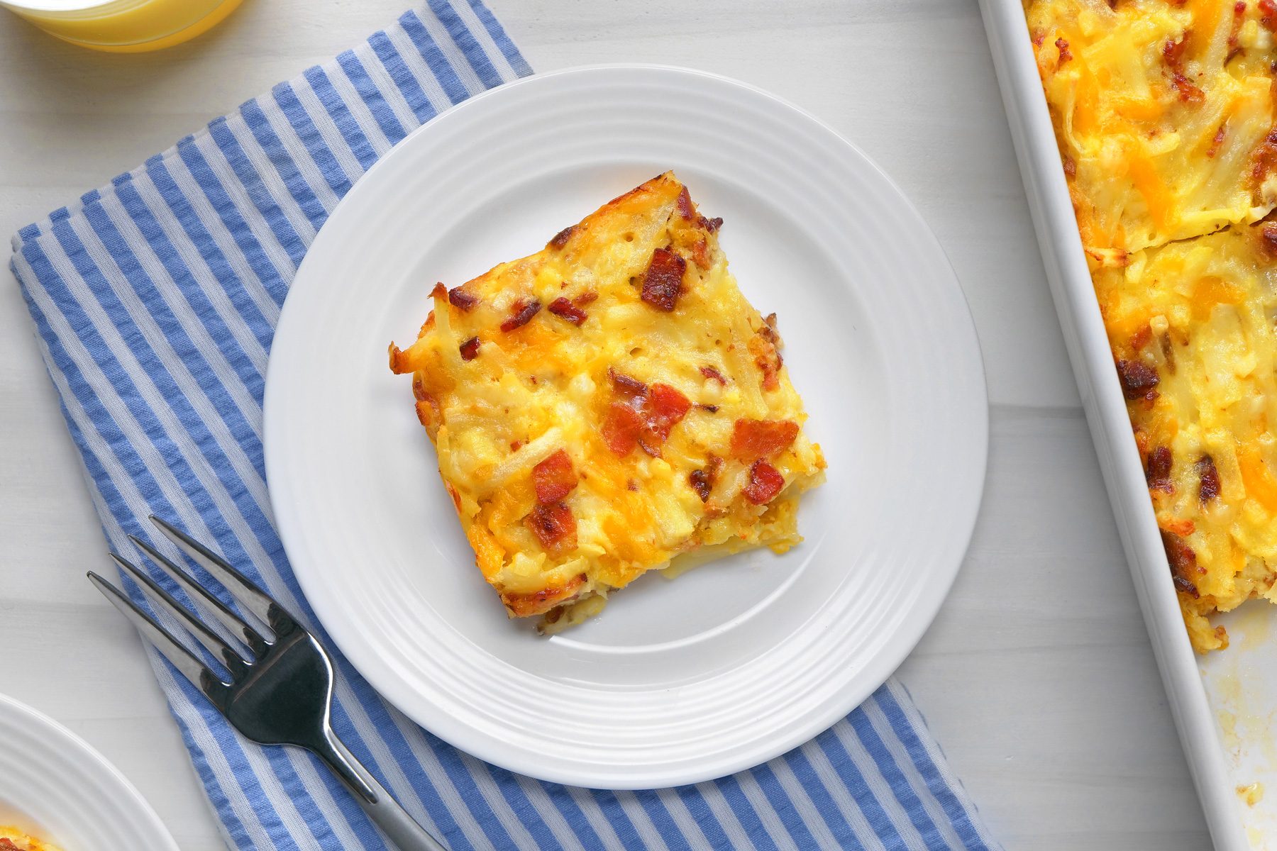 overhead shot; white background; Amish Breakfast Casserole in small white plate; Freshly cut squares are arranged on white plates beside the main dish; silver fork on blue and white striped napkin