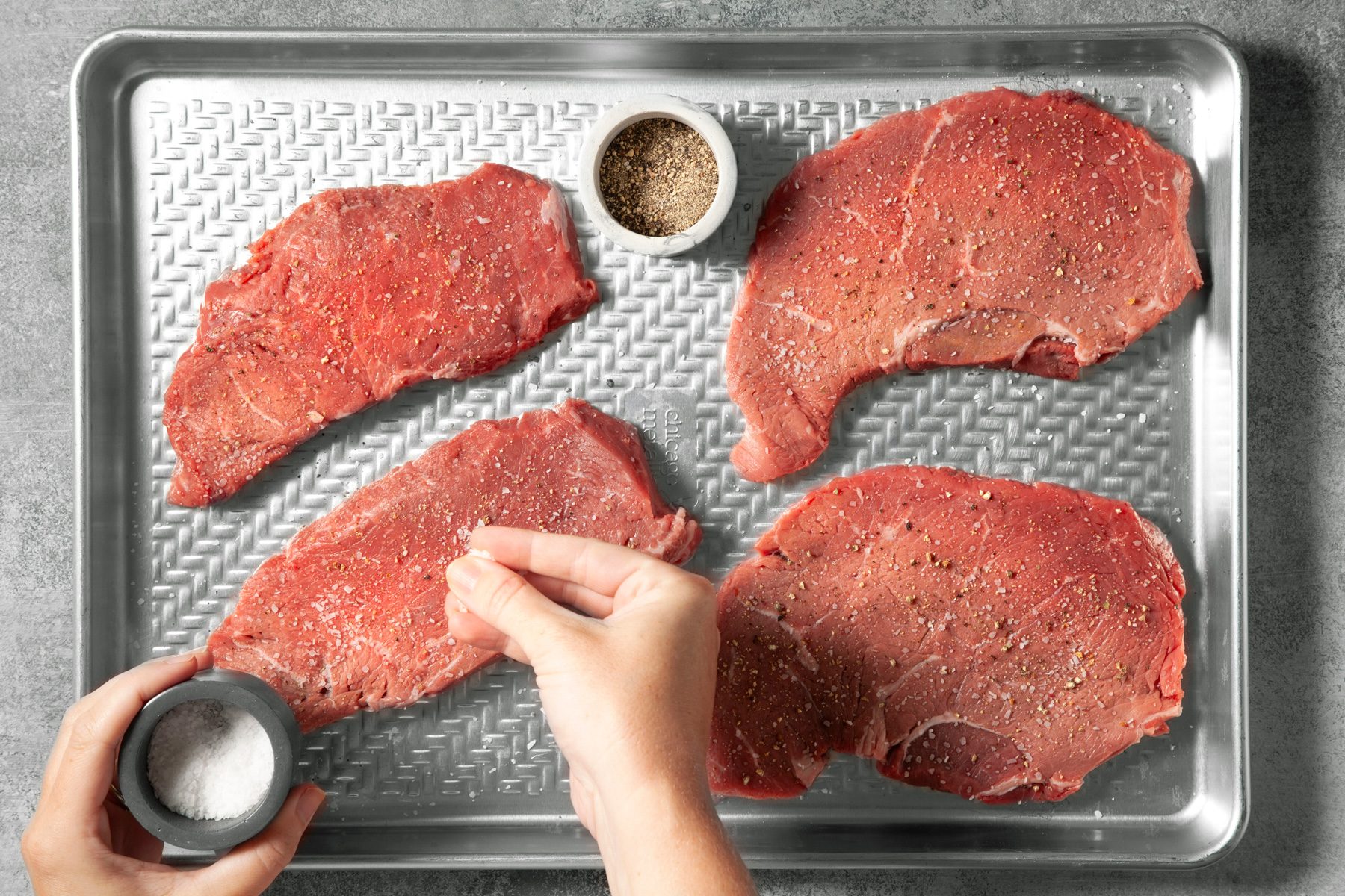 overhead shot; light grey background; Sprinkling steak with salt and pepper, placed over a silver baking tray