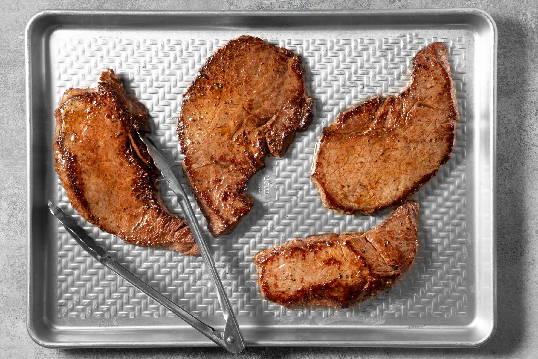 overhead shot; light grey background; transferred steak over baking tray, a tong is placed over the tray