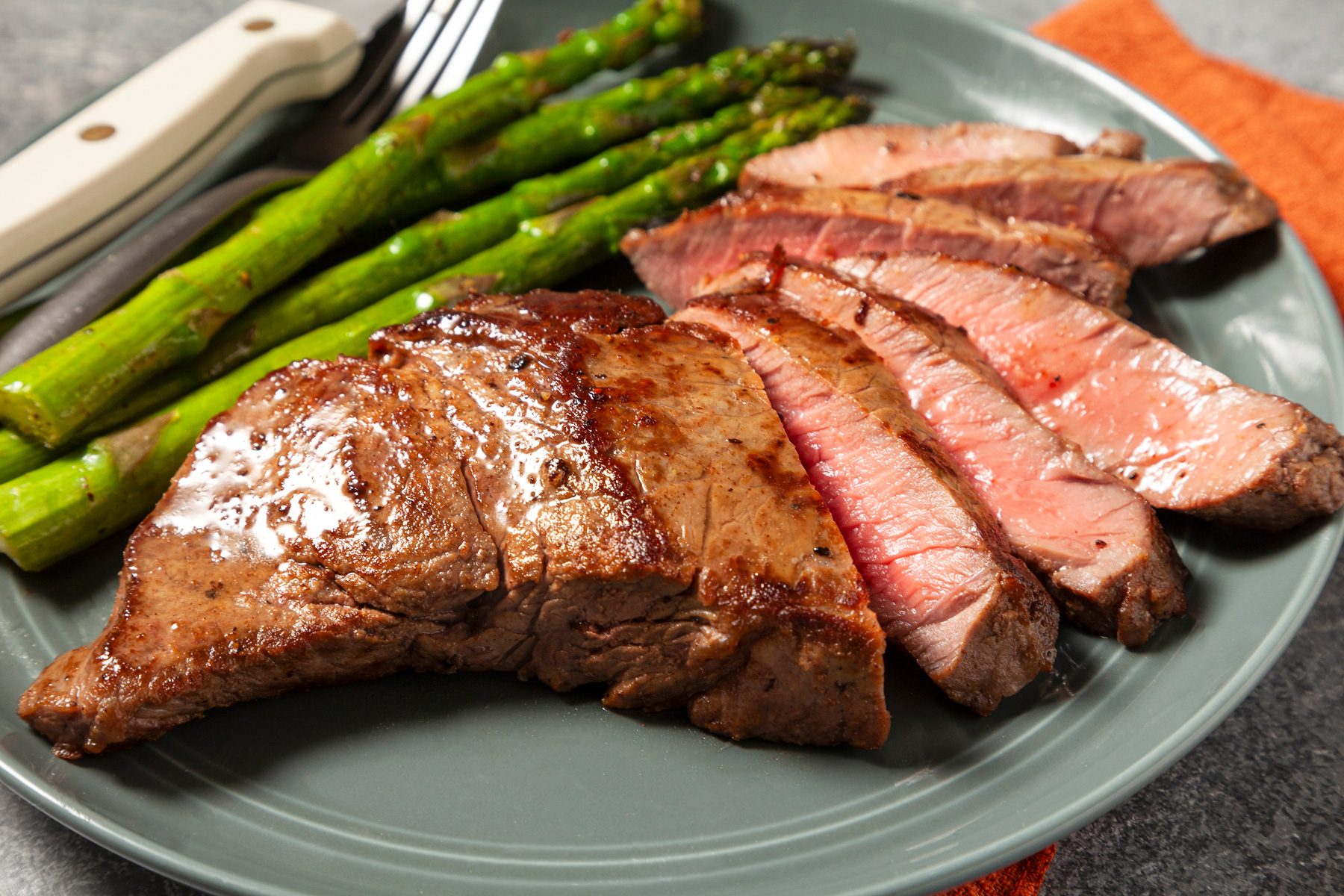 close shot; light grey background; a plate of sliced steak, takes center stage over red kitchen towel, accompanied by a side of roasted asparagus