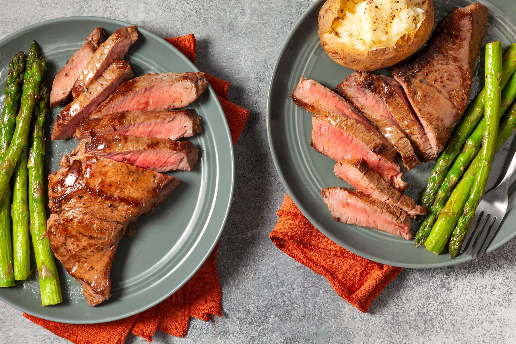 overhead shot; light grey background; two plates of sliced steak over red kitchen towel, accompanied by a side of roasted asparagus and a baked potato, with a silver spoon