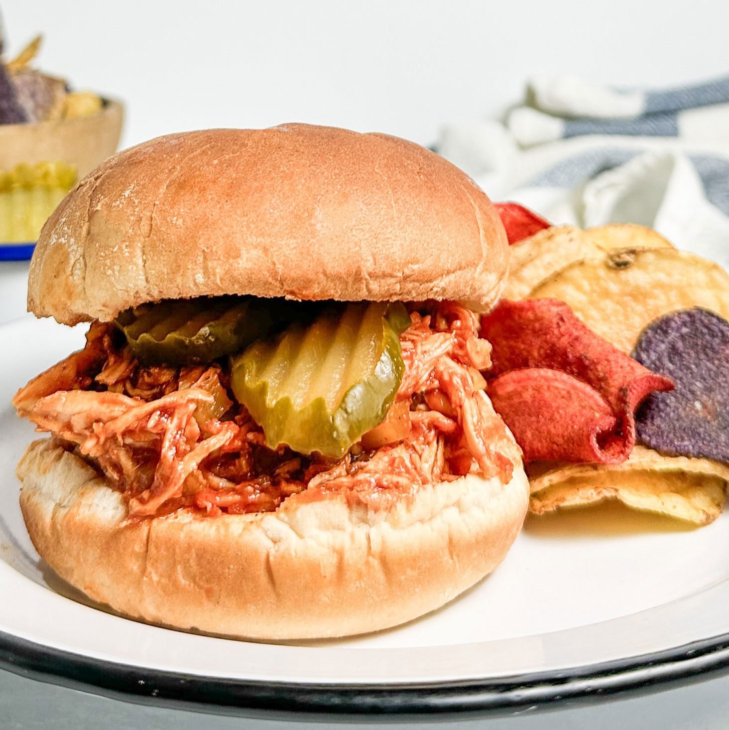 Taste of Home Barbecue Chicken Sandwich with chips on enamel plates next to bowls of chips and pickles