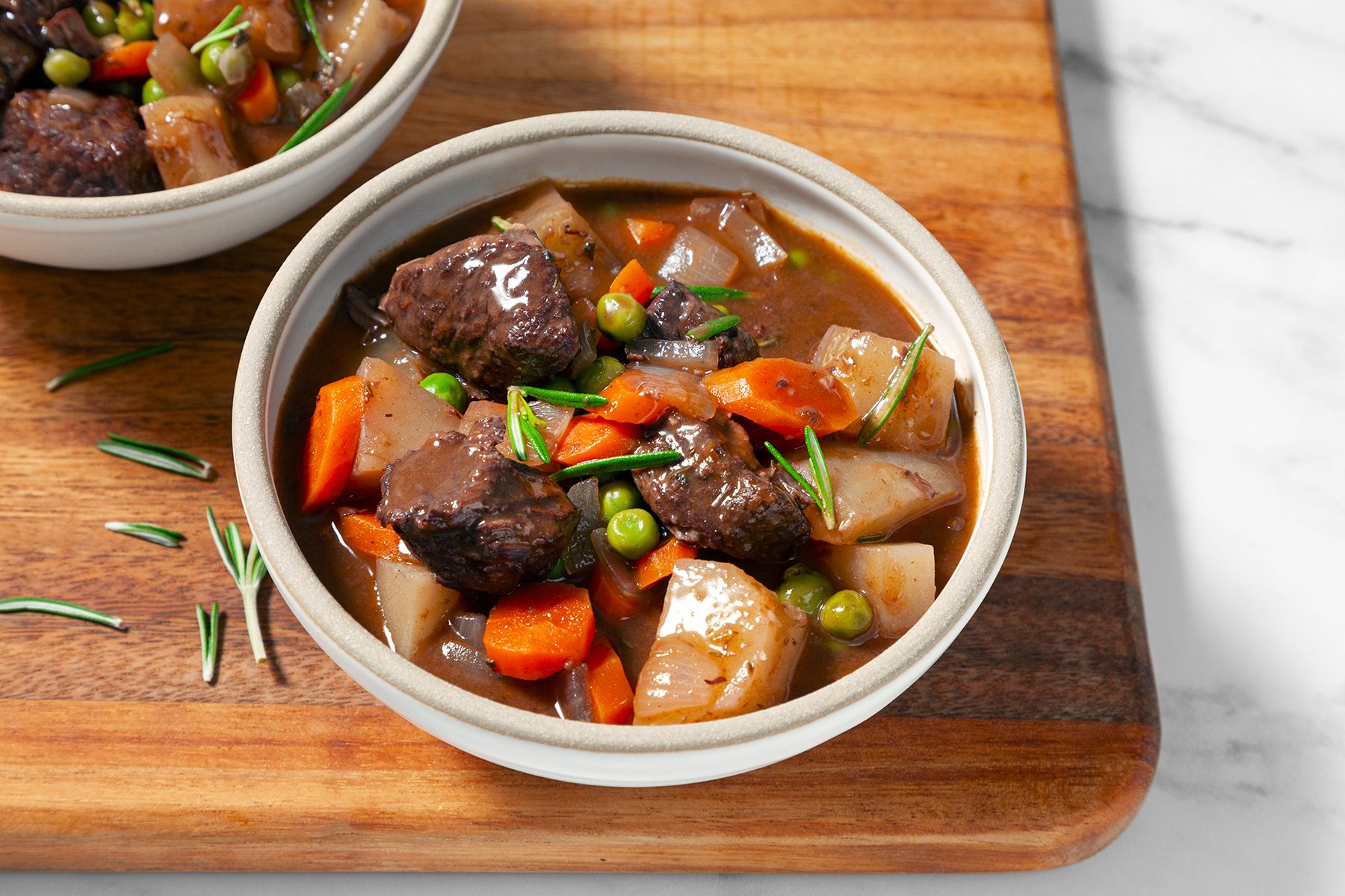 A bowl of hearty beef stew filled with chunks of tender beef, diced potatoes, carrots, peas, and onions, garnished with fresh rosemary. The bowl sits on a wooden cutting board with another identical bowl partially visible in the background.