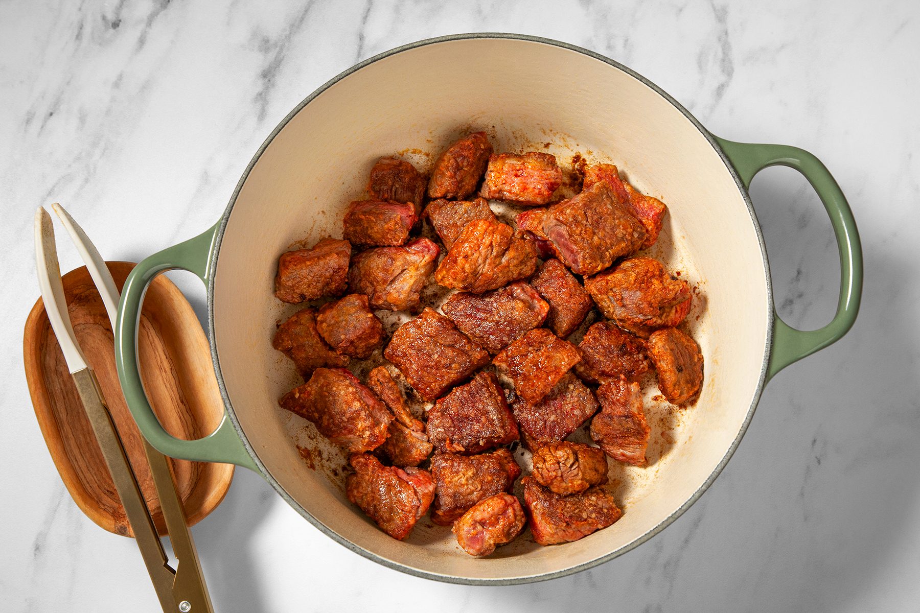 A green Dutch oven on a marble countertop containing seared beef chunks with a brown crust. Next to the pot is a wooden plate with silver tongs resting on it.