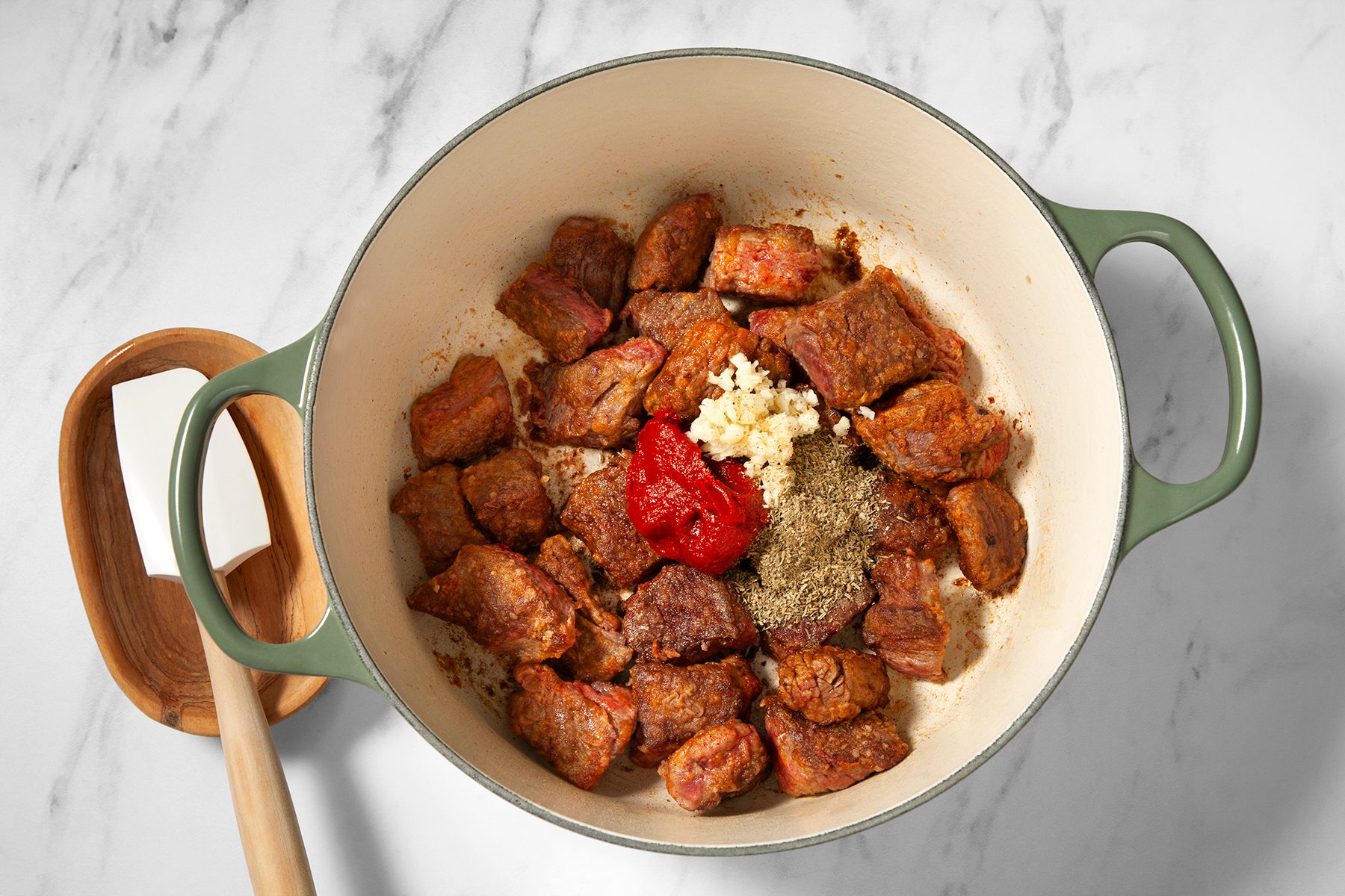 A green-handled pot placed on a white marble surface contains seared beef chunks, minced garlic, tomato paste, and herbs. A brown wooden spoon rest with a white spatula lies beside the pot.