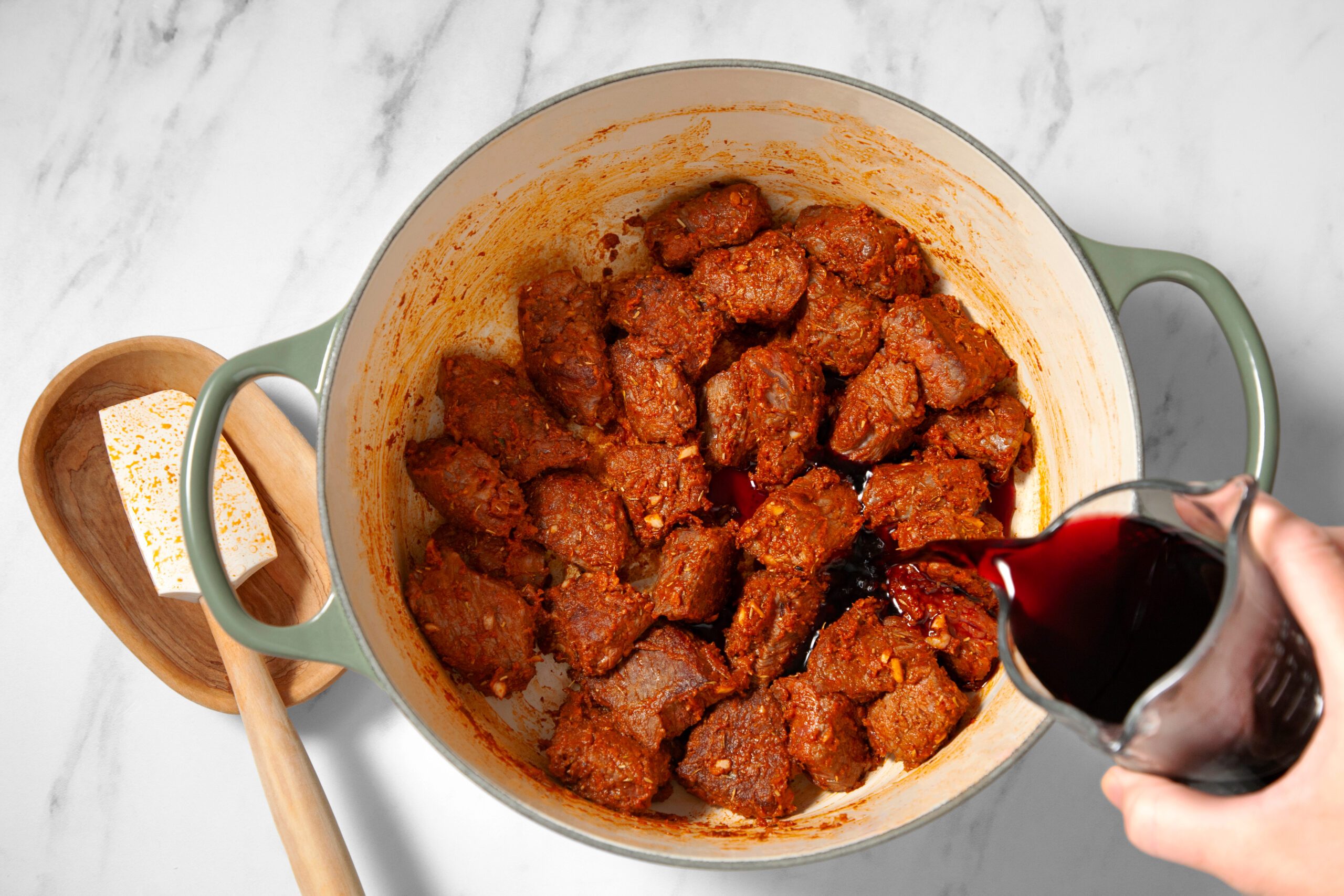 A person is pouring a dark liquid, possibly red wine, into a pot containing chunks of meat coated in spices, on a marble countertop. A wooden spoon rests by the side along with a small dish holding a block of butter.