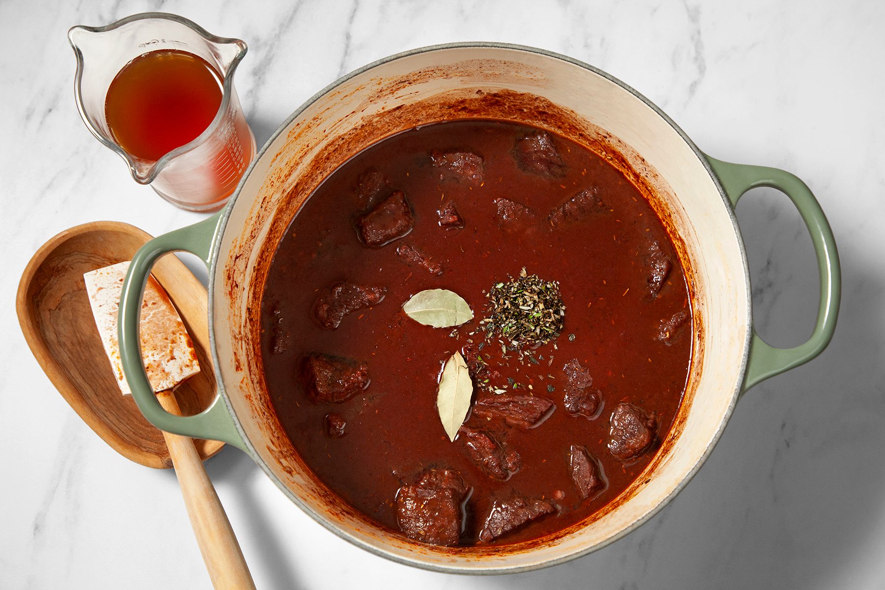 A large pot of rich, red stew containing chunks of meat, garnished with bay leaves and herbs, sits on a white marble countertop. A wooden spoon rests on a wooden holder to the side. A small pitcher filled with broth or sauce is nearby.