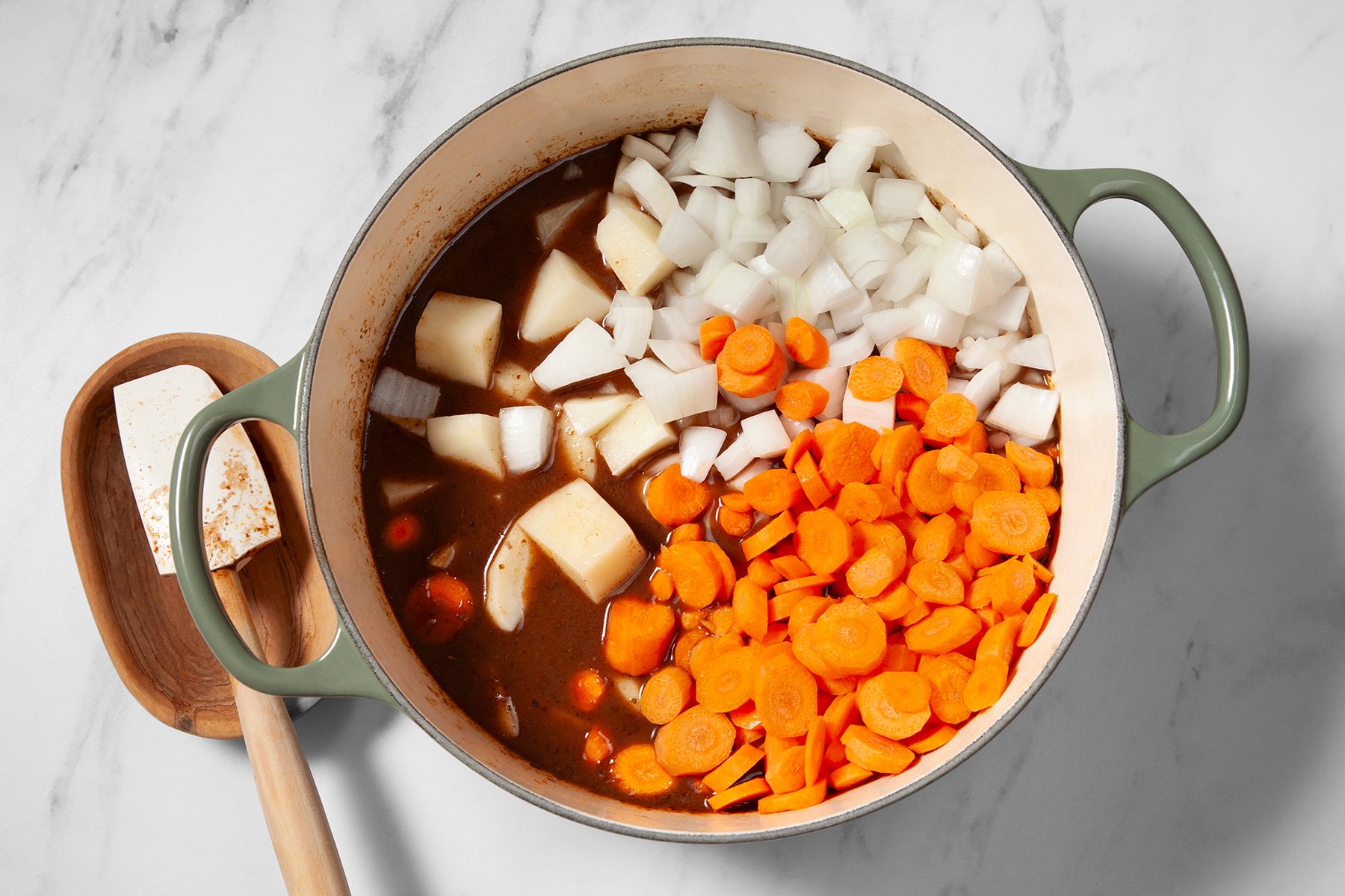 A green-handled pot on a marble countertop contains a stew with large chunks of onions, carrots, and potatoes in a brown broth. A wooden spoon rests on a small wooden tray, beside the pot. The vegetables appear to be in the process of being cooked.