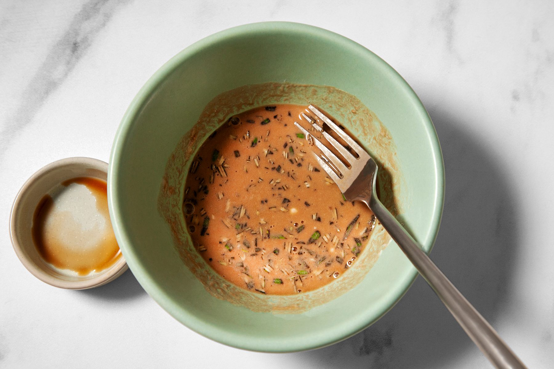 A green bowl containing a creamy, seasoned mixture with herbs, and a fork resting inside. Next to the bowl, there is a small lid with some residue on it, all set on a white marble surface.