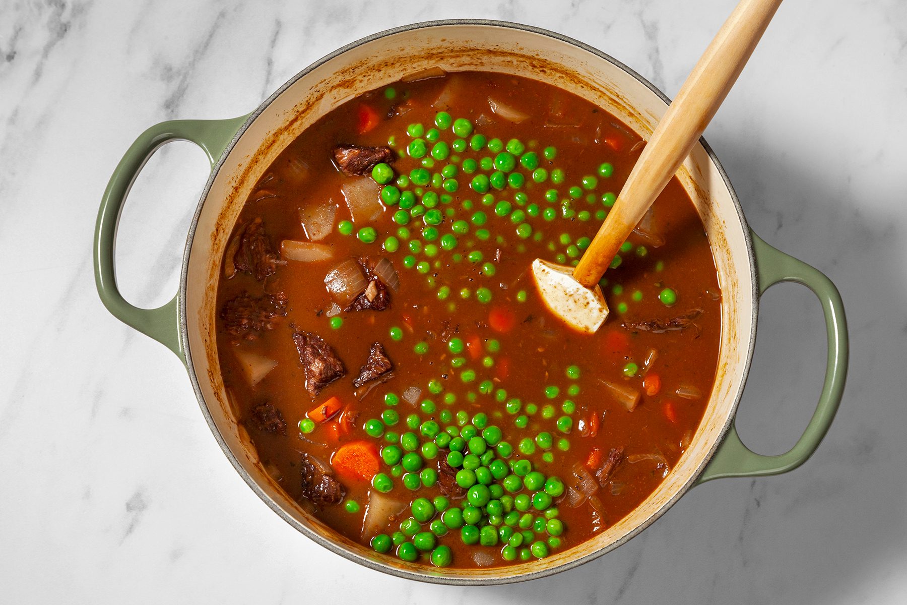 A green pot filled with a rich, brown beef stew containing chunks of meat, diced potatoes, carrots, and peas, sits on a white marble surface. A wooden spoon rests in the stew.