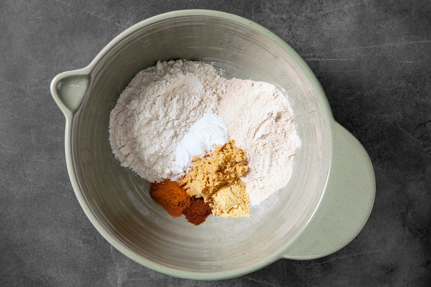 A mixing bowl on a dark countertop contains several dry ingredients: flour, baking soda, ground ginger, cinnamon, and clove powder. The ingredients are not yet mixed, creating distinct sections of different colors and textures in the bowl.