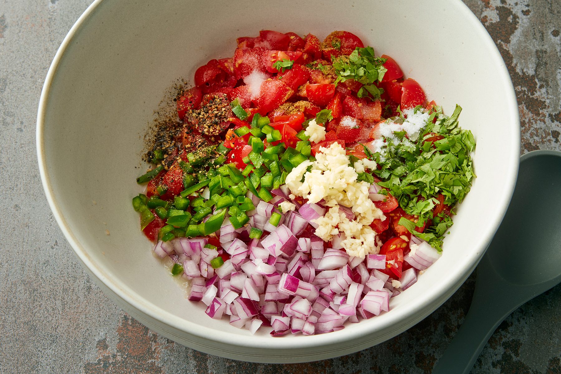 A large white bowl contains chopped red onions, minced garlic, diced green bell peppers, chopped tomatoes, chopped cilantro, and assorted spices. A grey serving spoon is placed beside the bowl on a textured surface.