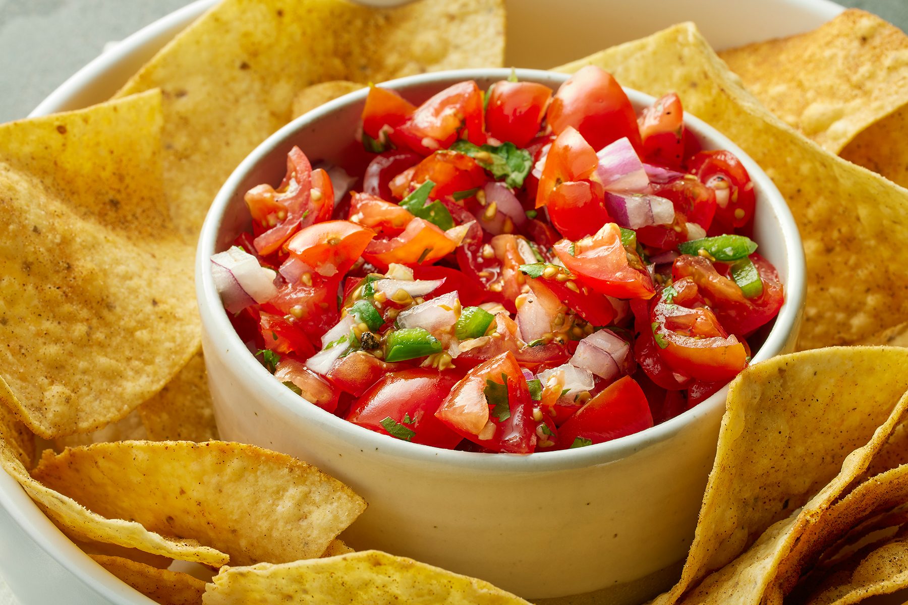 A bowl of fresh tomato salsa placed in the center of a dish surrounded by crispy tortilla chips. The salsa contains diced tomatoes, onions, and herbs, creating a colorful, appetizing contrast with the golden-brown chips.