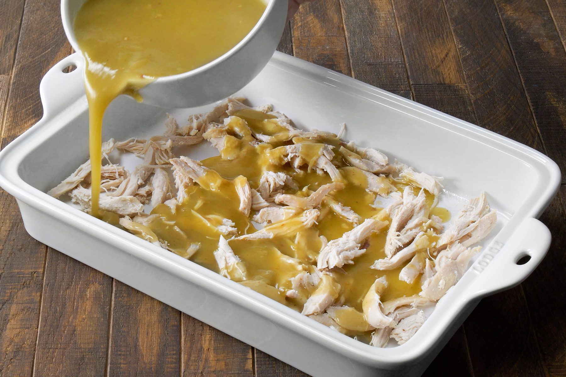 3/4th shot; wooden background; A golden-brown sauce is being poured over shredded chicken in a white baking dish, The chicken is pale white and the sauce is thick and creamy