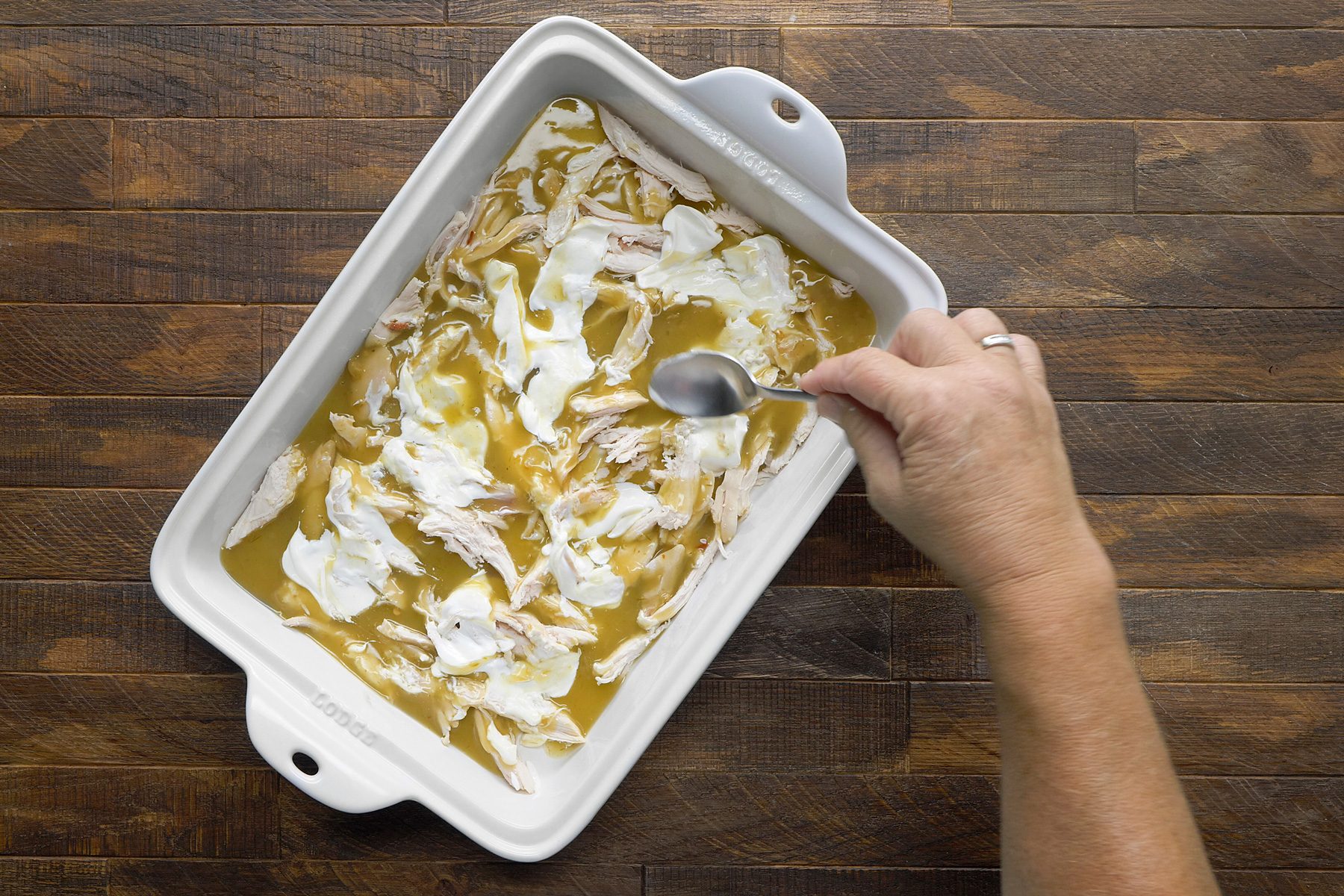 overhead shot; wooden background; A white baking dish contains shredded chicken and a golden-brown sauce, A person is using a spoon to stir the mixture together, ensuring the chicken is evenly coated in the sauce, The sauce appears thick and creamy, while the chicken is pale white