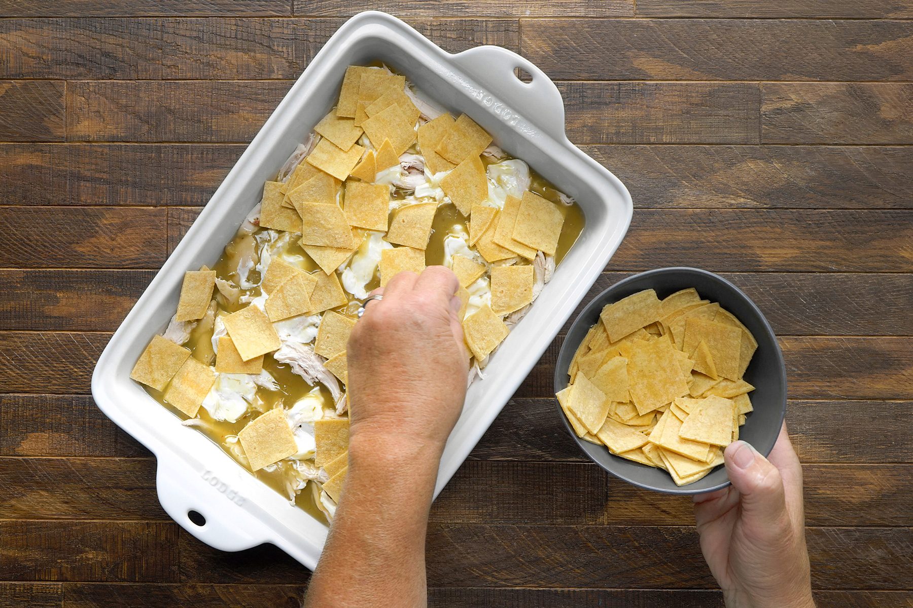 overhead shot; wooden background; A person is adding tortilla chips to a baking dish filled with shredded chicken and a creamy sauce, The tortilla chips are being arranged in a single layer on top of the chicken mixture, A bowl of additional tortilla chips is visible on the side