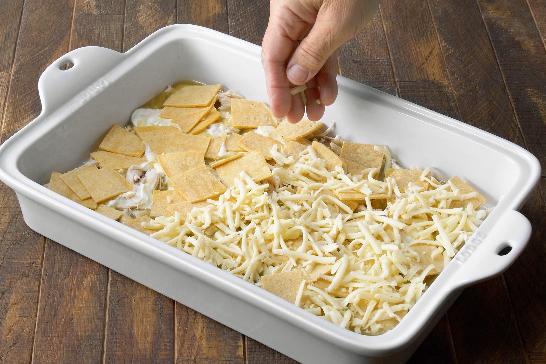 3/4th shot; wooden background; A white baking dish contains a layer of tortilla chips, a layer of shredded chicken, and a layer of creamy sauce, A person is sprinkling shredded cheese on top of the dish, The cheese is a pale yellow color and is covering the entire surface of the dish