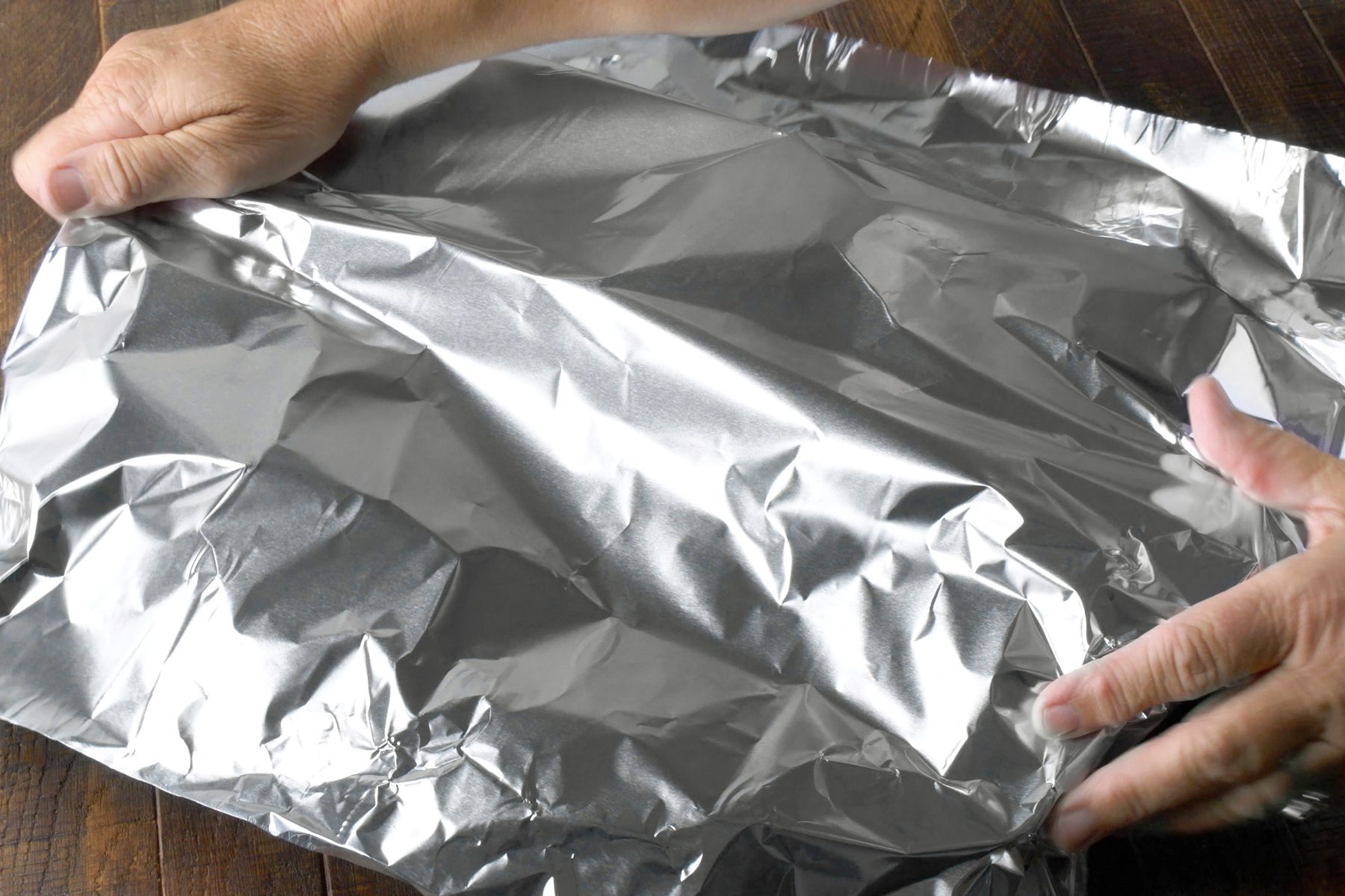 3/4th shot; wooden background; A person is folding aluminum foil over a baking dish, The foil is being wrapped tightly around the dish to create a sealed tent, The baking dish is not visible, but the foil is forming a large, rectangular shape