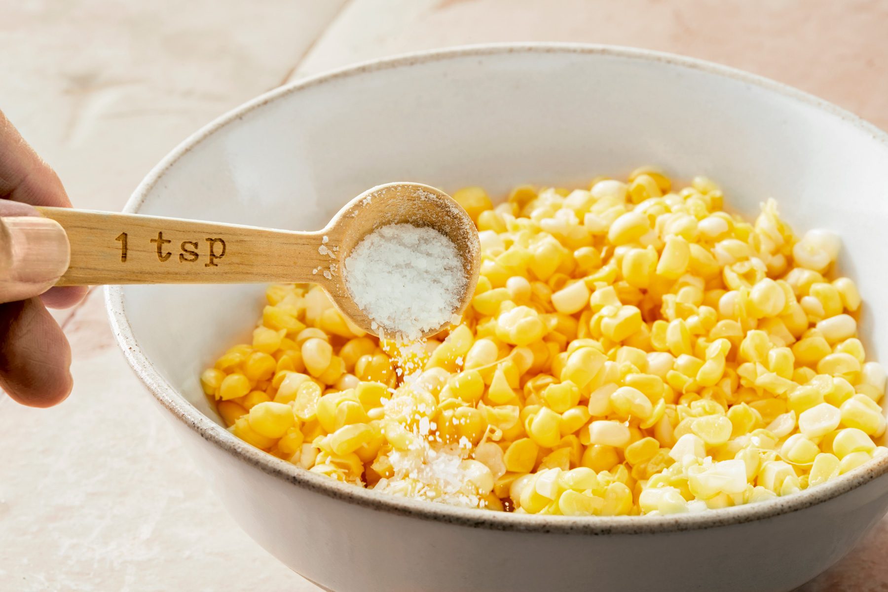 Table view shot of a small bowl; combine corn and salt; Let sit 10 minutes; wooden table spoon; cream square tiles background;