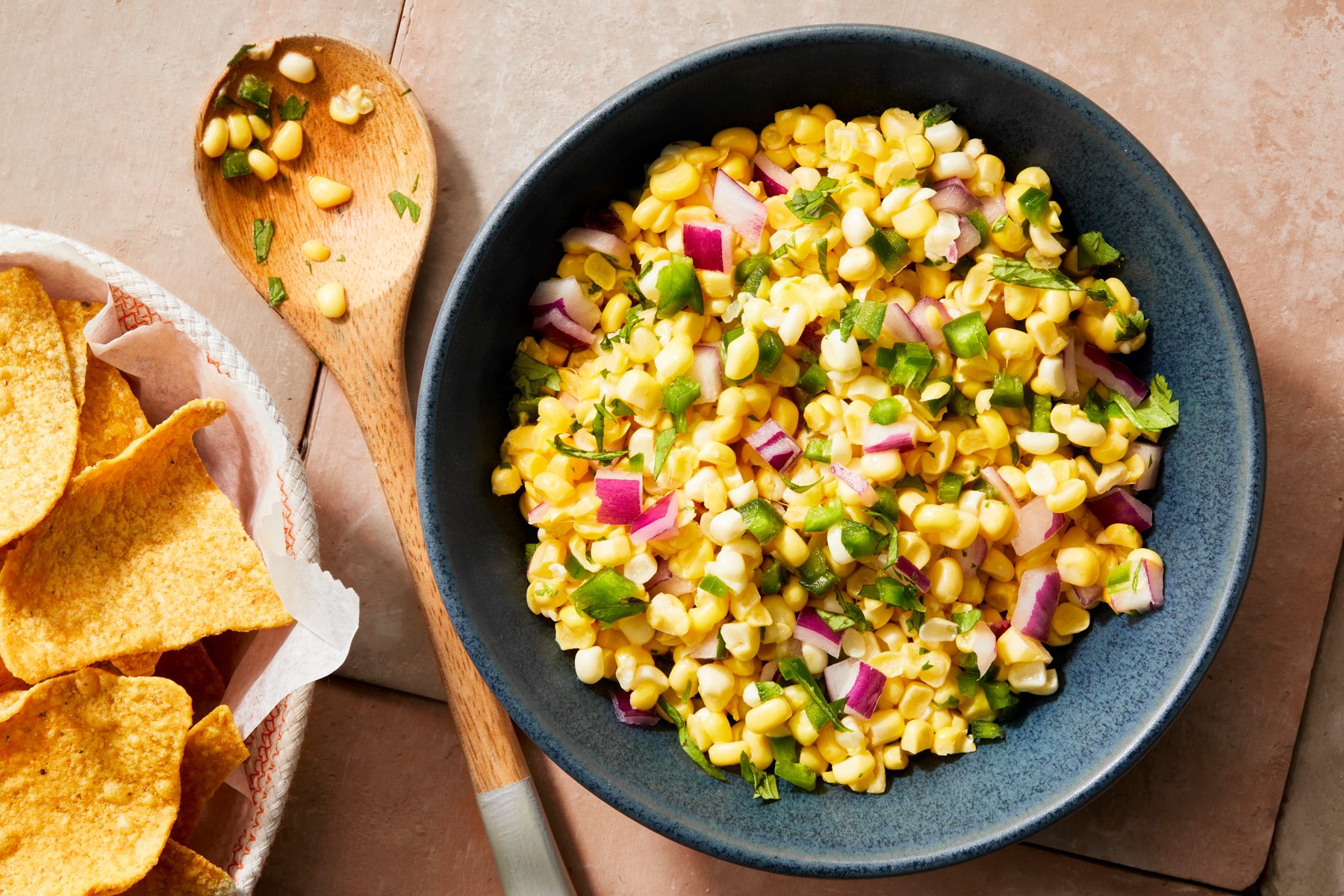 Overhead shot of Copycat Chipotle Corn Salsa; in a black bowl; serve with fresh corn chips bowl; wooden spoon; cream square tiles background