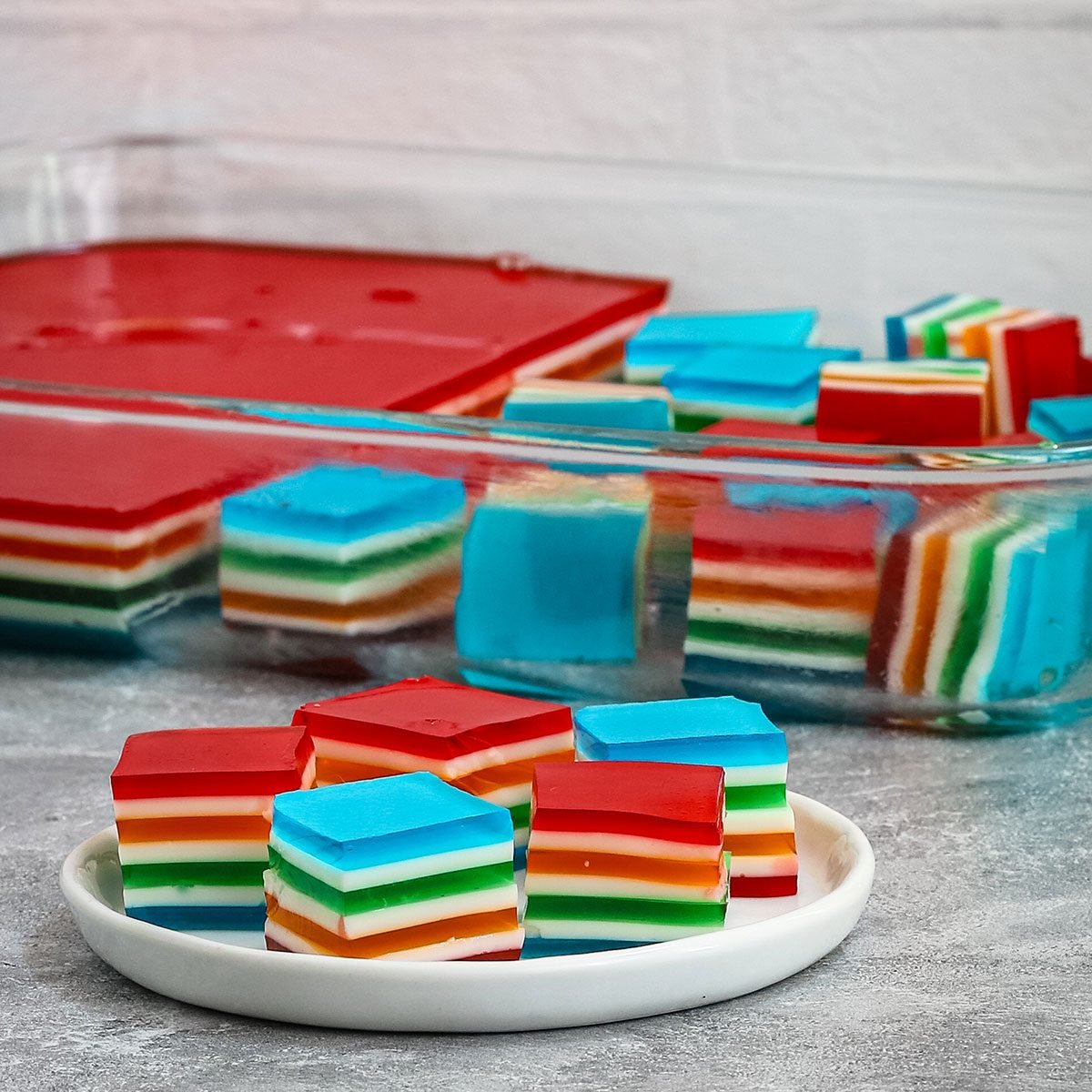 Rainbow jello cubes on a small plate with more in the background.