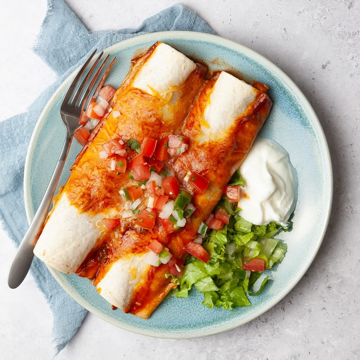 Single serving of Taste of Home Crock-Pot Chicken Enchiladas garnished with pice de gallo, lettuce and sour cream. Small bowls of pico and lettuce.