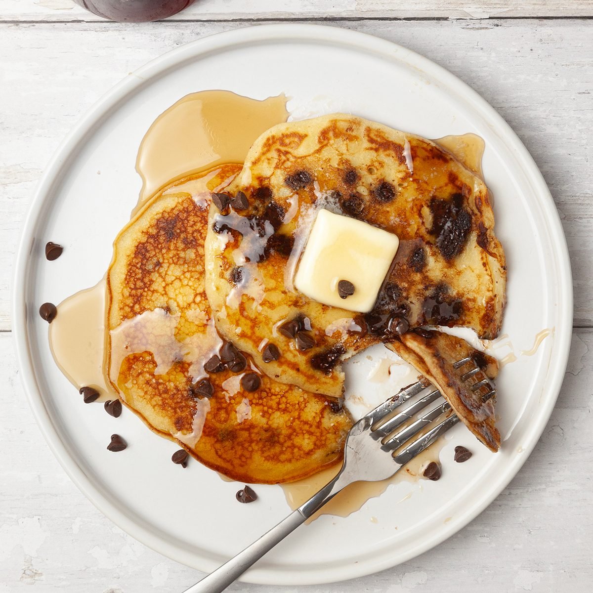 Single serving of Taste of Home Chocolate Chip Pancakes on a white platter, plate for butter pats, syrup cruet, mini chocolate chips