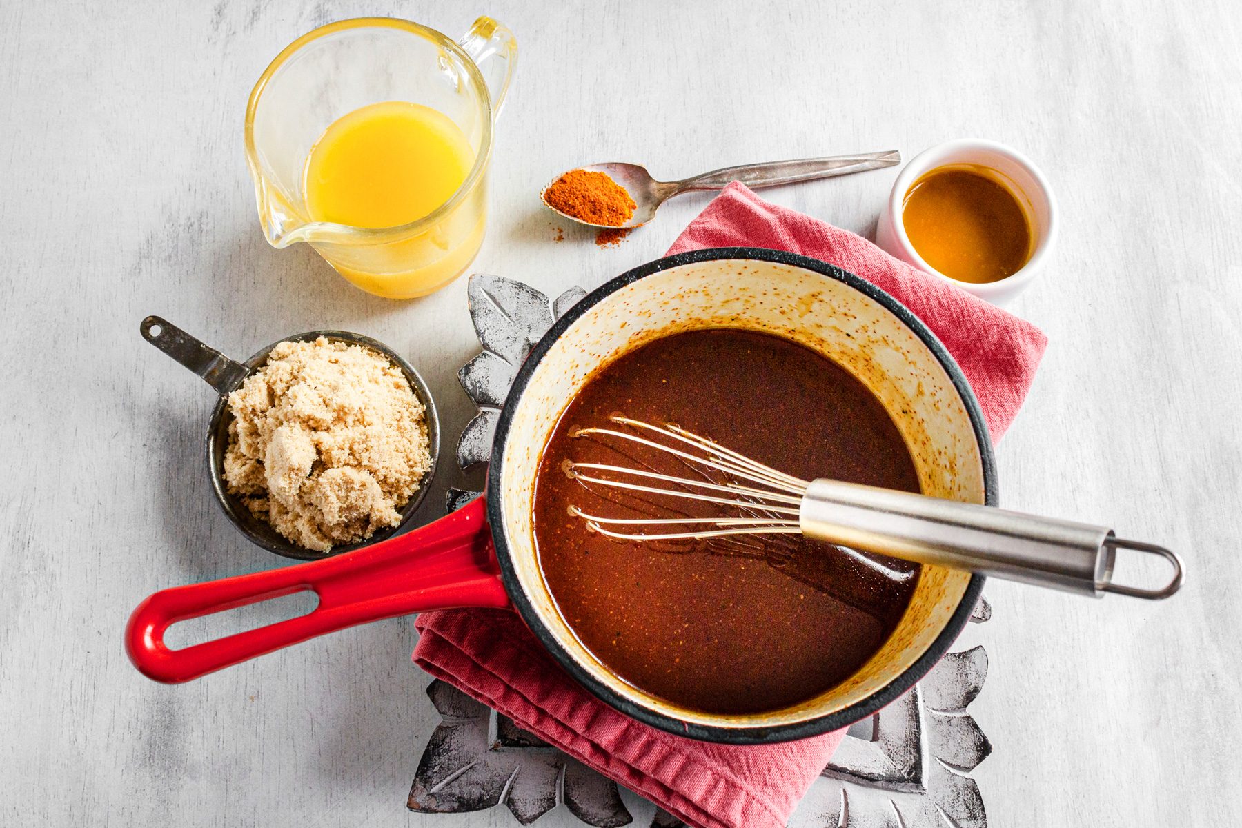 overhead shot; white background; glaze mixture in saucepan; whisker; brown sugar in measuring cup; pineapple juice in glas beaker; pepperr in spoon; honey; kitchen towel;