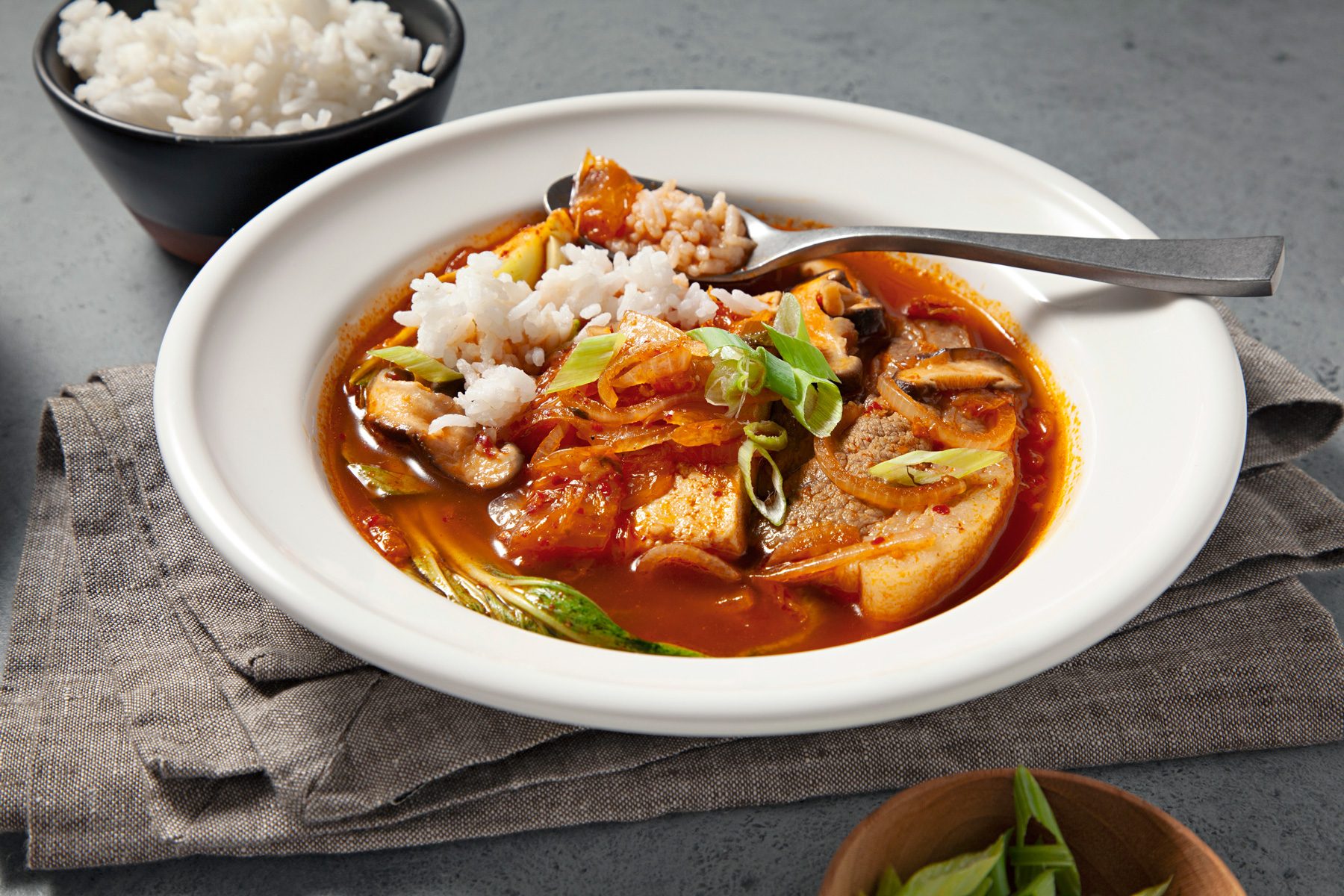 3/4th shot; light grey background; a bowl of Kimchi Soup is presented, showcasing a vibrant red broth filled with chunks of tofu, kimchi, and other vegetables; A side of white rice is served alongside the soup, Chopped green onions garnish the soup