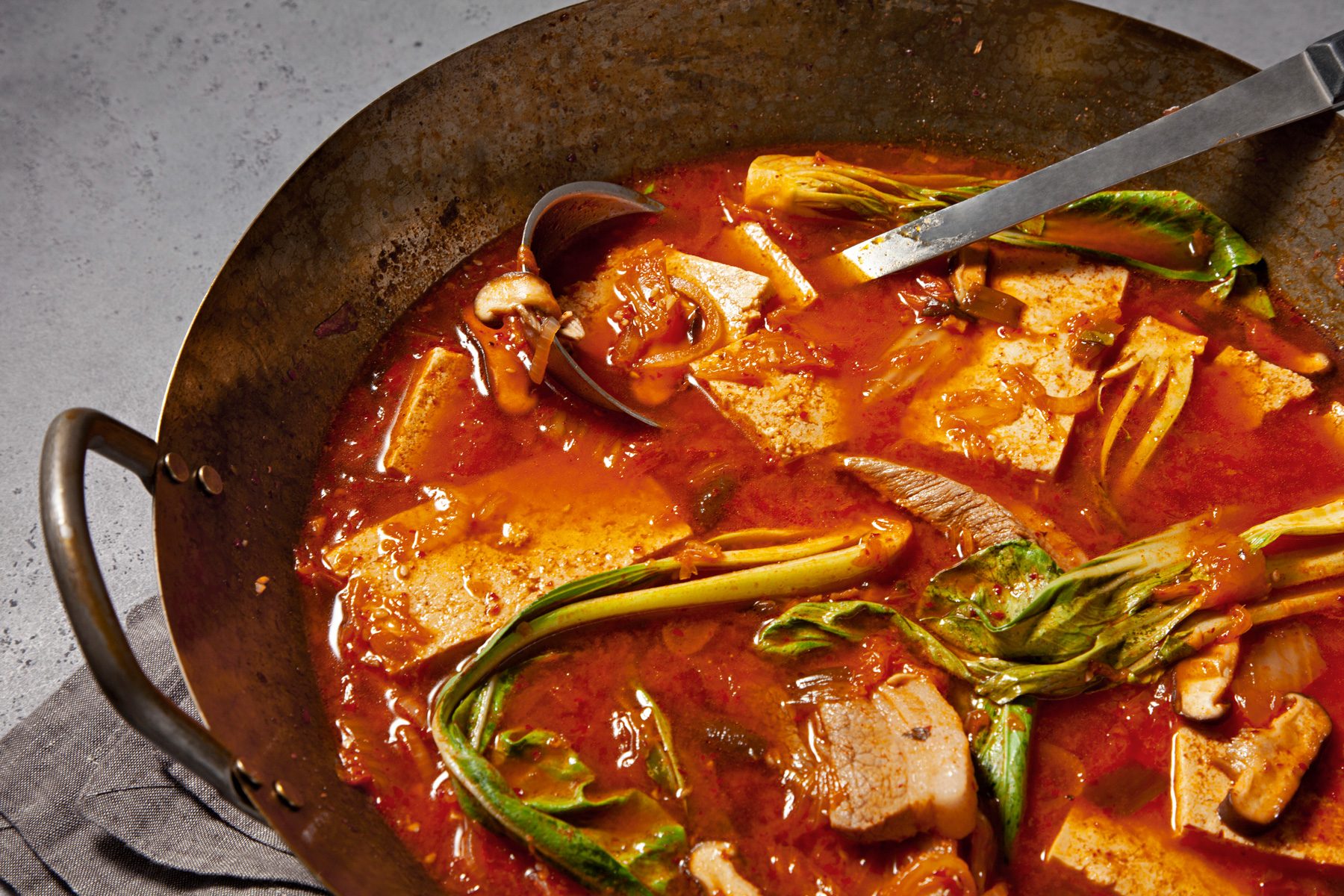 3/4th shot; light grey background; A steaming bowl of Kimchi Soup is the centerpiece of the image; The soup is a vibrant red color, filled with chunks of tofu, kimchi, and other vegetables; A ladle rests in the bowl, ready to serve the flavorful broth