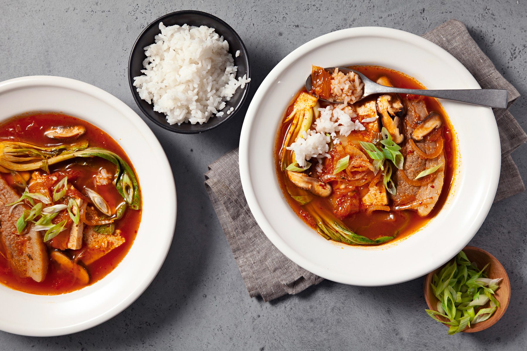 overhead shot; light grey background; Two steaming bowls of Kimchi Soup are presented, showcasing a vibrant red broth filled with chunks of tofu, kimchi, and other vegetables; A side of white rice is served alongside the soup, Chopped green onions garnish the soup