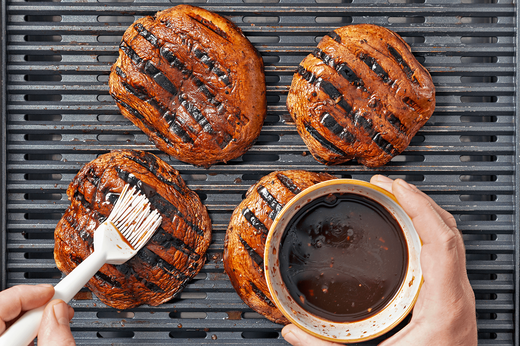 A pair of hands is shown grilling four portobello mushroom caps on a grill. One hand holds a bowl of dark glaze or marinade, while the other hand uses a brush to apply the glaze to one of the mushrooms. The mushrooms have distinct grill marks.