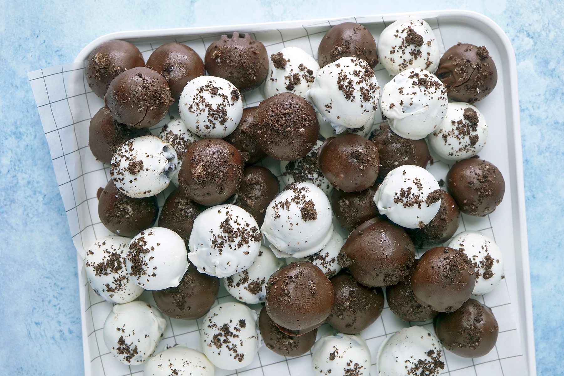 A tray filled with an assortment of chocolate truffles, some coated in white chocolate and others in dark chocolate. The truffles are sprinkled with cookie crumbs and arranged on a checkered paper-lined tray, placed on a light blue surface.