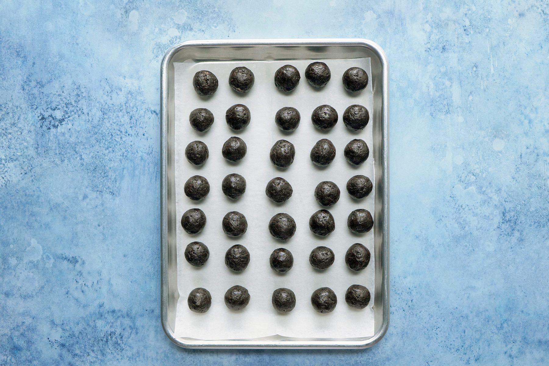 A baking sheet lined with parchment paper holds five rows of chocolate truffles, with each row containing seven round truffles. The background is a textured, light blue surface.