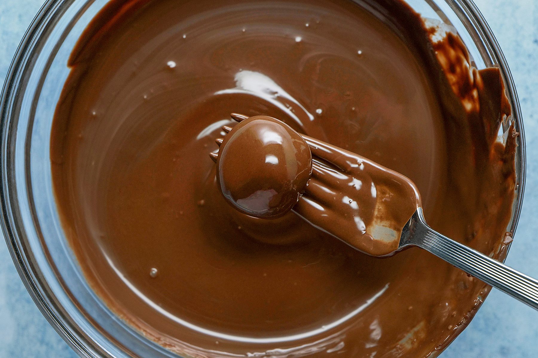 A metal fork holding a chocolate-coated treat is partially submerged in a bowl of melted chocolate. The smooth, glossy surface of the chocolate covers both the treat and the fork, indicating that it has just been dipped. 