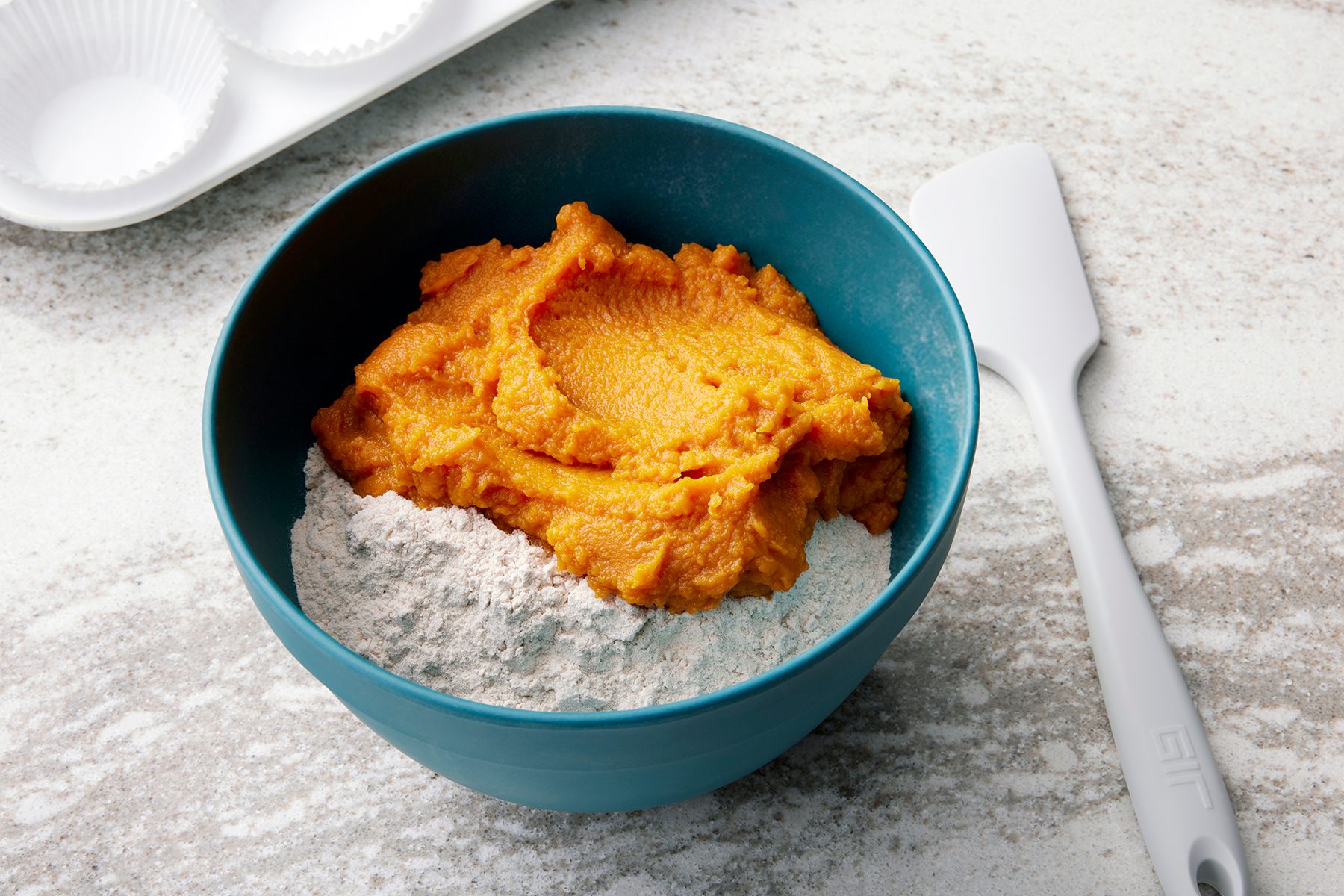 A blue bowl containing a mixture of pumpkin puree and flour sits on a light-colored countertop. A white spatula is placed beside the bowl. A cupcake tray with paper liners is partially visible in the background.