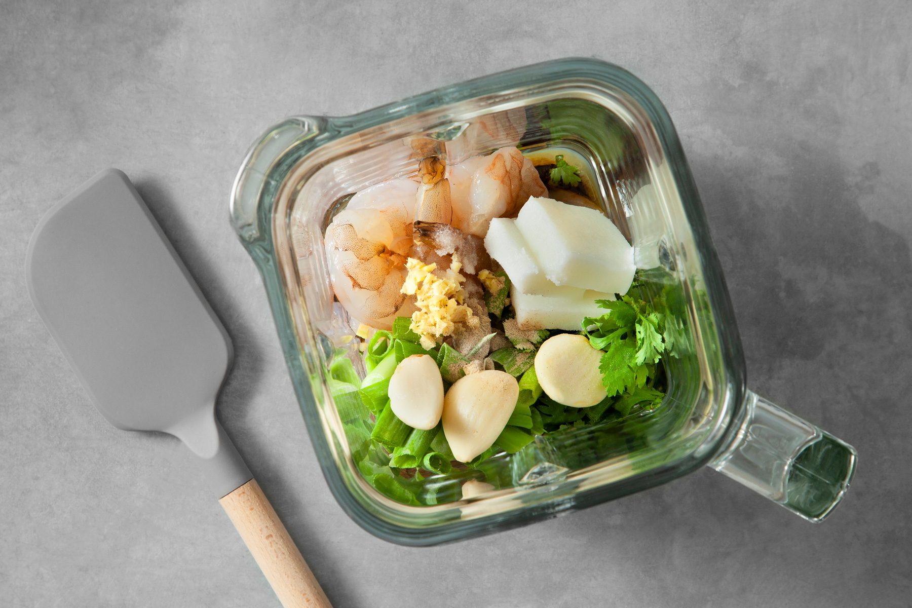 overhead shot; light grey background; A blender containing a vibrant mixture of ingredients for a Shrimp Toast recipe; Peeled and deveined shrimp, ginger, garlic, and green onions; a spatula is lying next to the blender