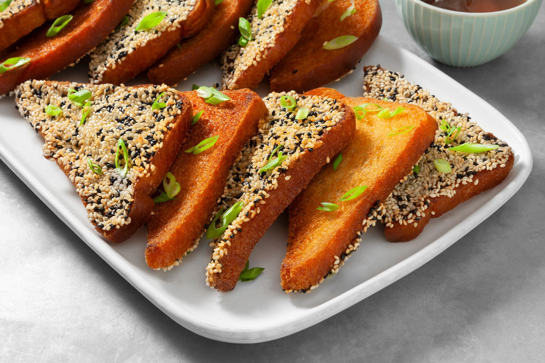 3/4th shot; close shot; light grey background; A platter of crispy shrimp toast is presented; The toast is golden brown and coated in a mixture of sesame seeds; Sliced green onions are sprinkled on top; A small bowl of dipping sauce accompanies the toast