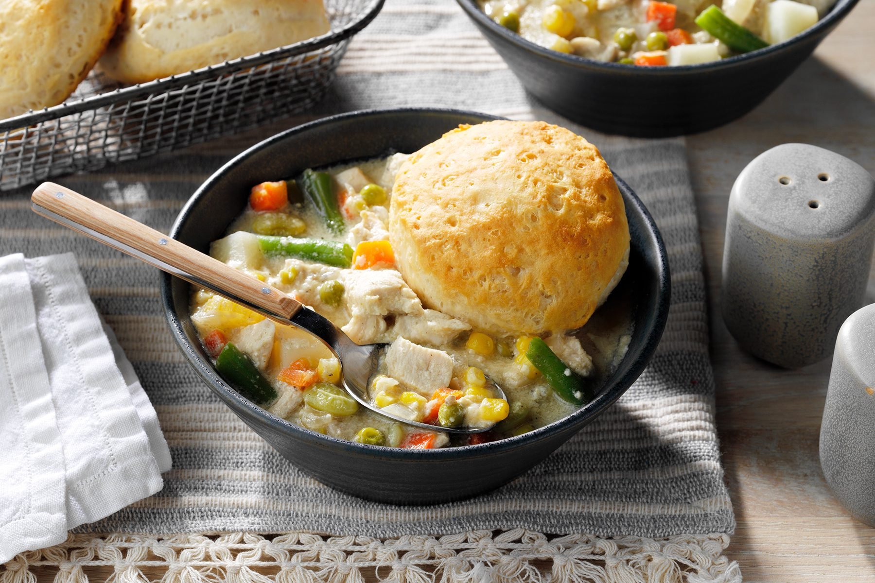 A black bowl filled with creamy chicken pot pie soup, featuring chunks of chicken, green beans, peas, carrots, corn, and potatoes. A golden-brown biscuit rests on top of the soup. In the background, a wire basket holds more biscuits on a striped cloth.