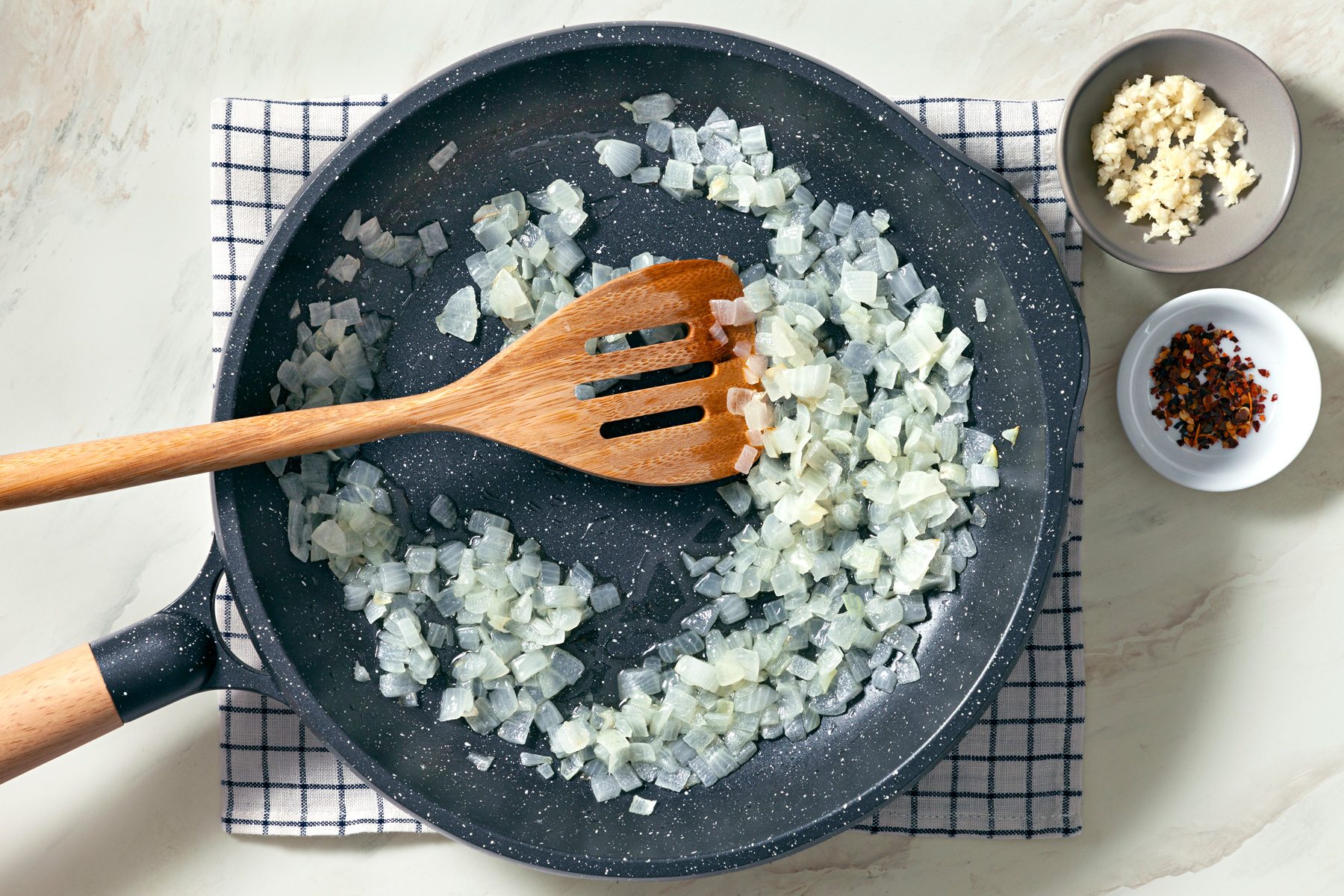 Overhead shot of a large skillet; heat oil over medium heat; add onion; cook and stir until tender; add garlic and pepper flakes; cook 1 minute longer; wooden flat spoon; kitchen towel; bowls of chilli flakes and garlic; marbel surface