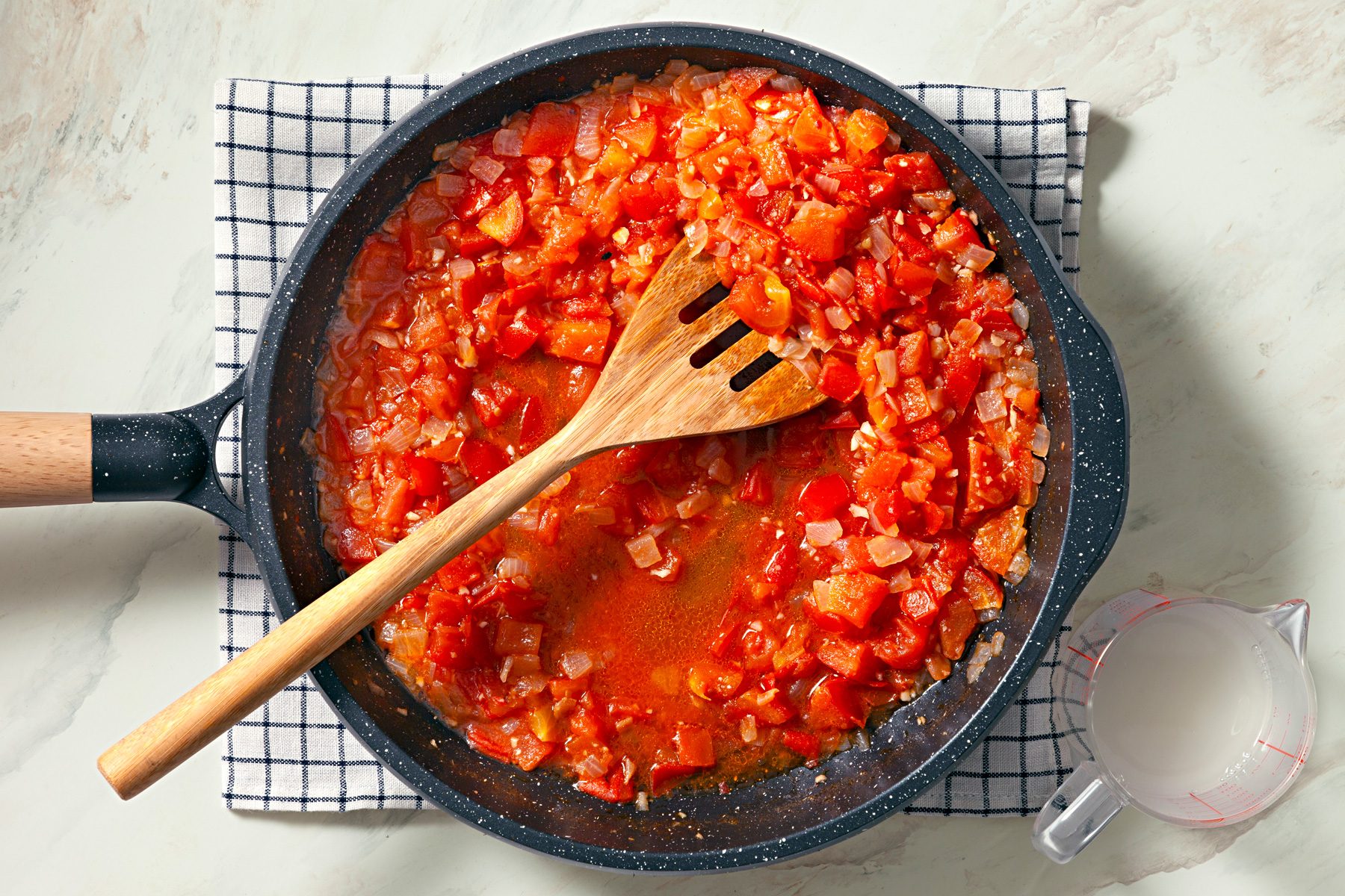 Overhead shot of add tomatoes and reserved pasta water to skillet; bring to a boil; reduce heat; simmer uncovered until sauce begins to thicken; kitchen towel; wooden flat spoon; marble surface