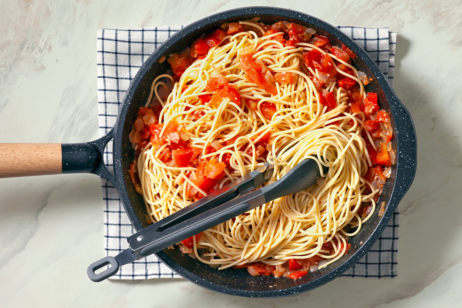 Overhead shot of add spaghetti and toss to coat; kitchen towel; tongs; marble surface