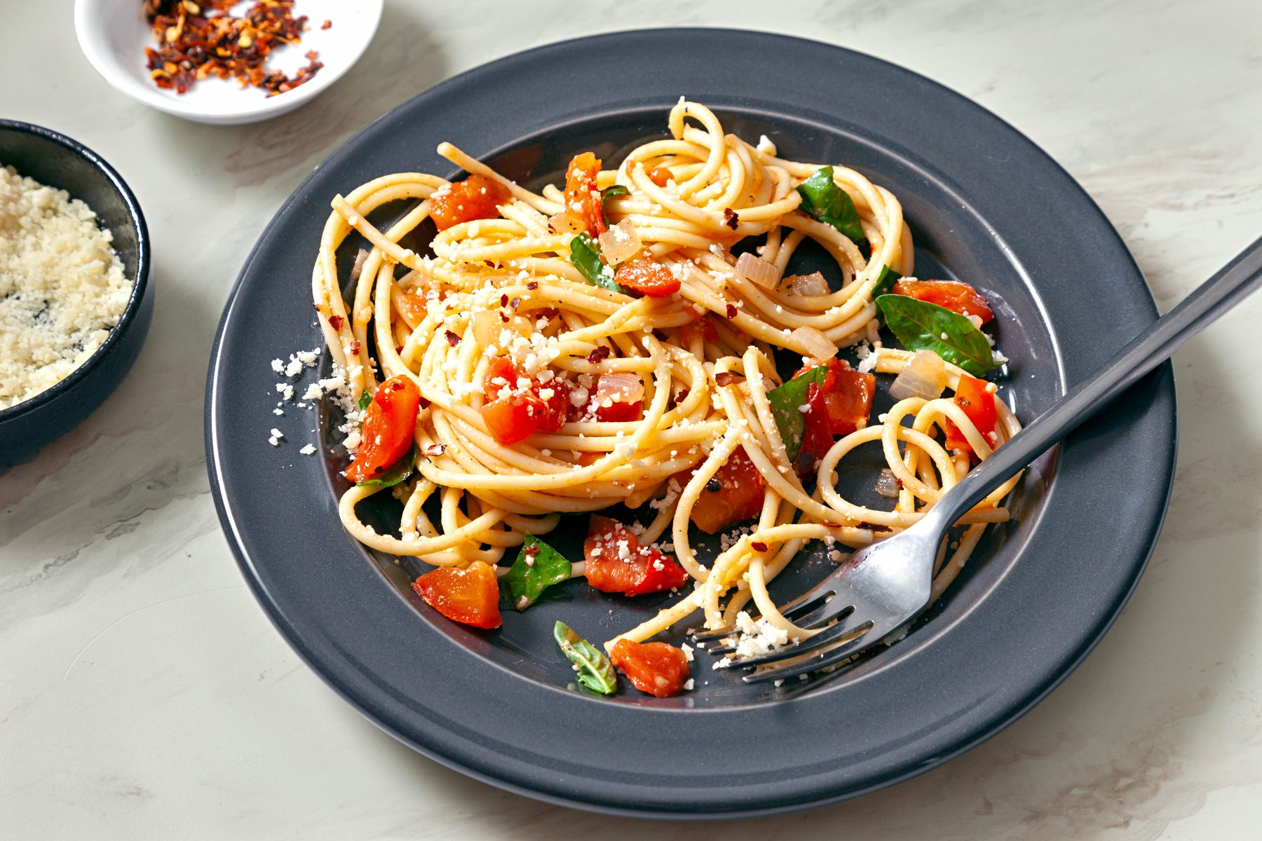 3/4 angle view shot of Spaghetti Pomodoro; serve on plate with Parmesan cheese; and top with additional fresh basil and crushed red pepper flakes; fork; marble surface