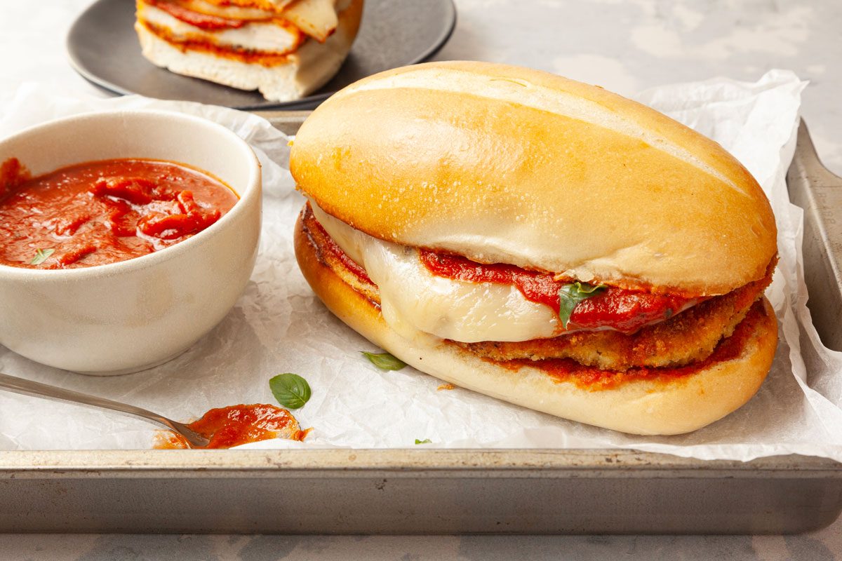 Two Taste of Home Parmesan Chicken Sandwiches with a bowl of marinara sauce on a metal tray