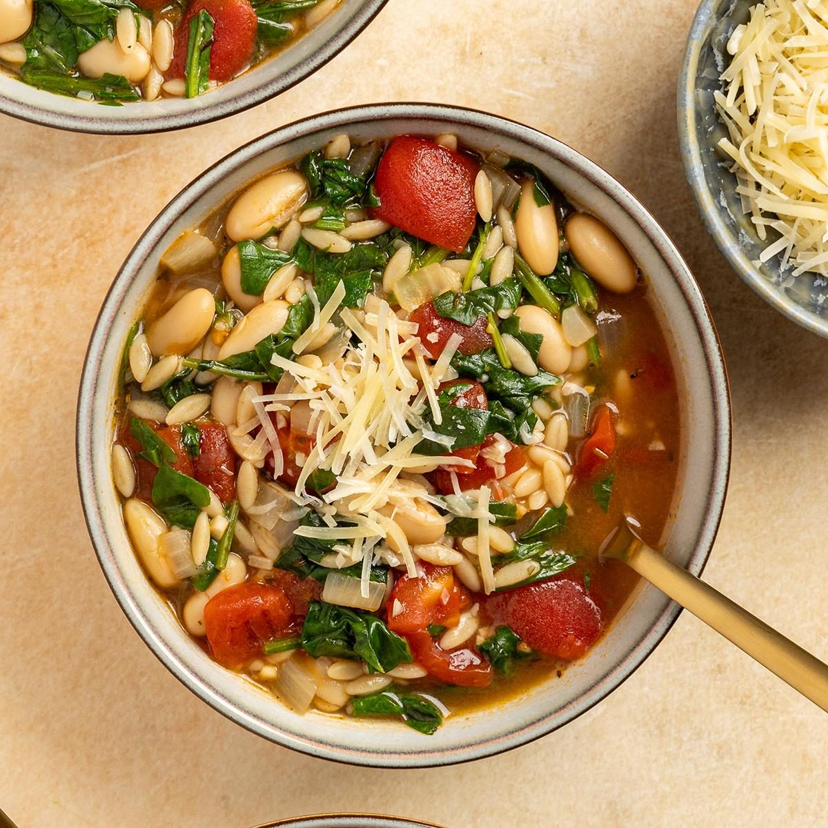 A bowl of soup containing orzo pasta, white beans, spinach, diced tomatoes, and grated cheese on top. The soup is placed on a light-colored surface, and a gold spoon rests inside the bowl. Another bowl of cheese is partially visible nearby.