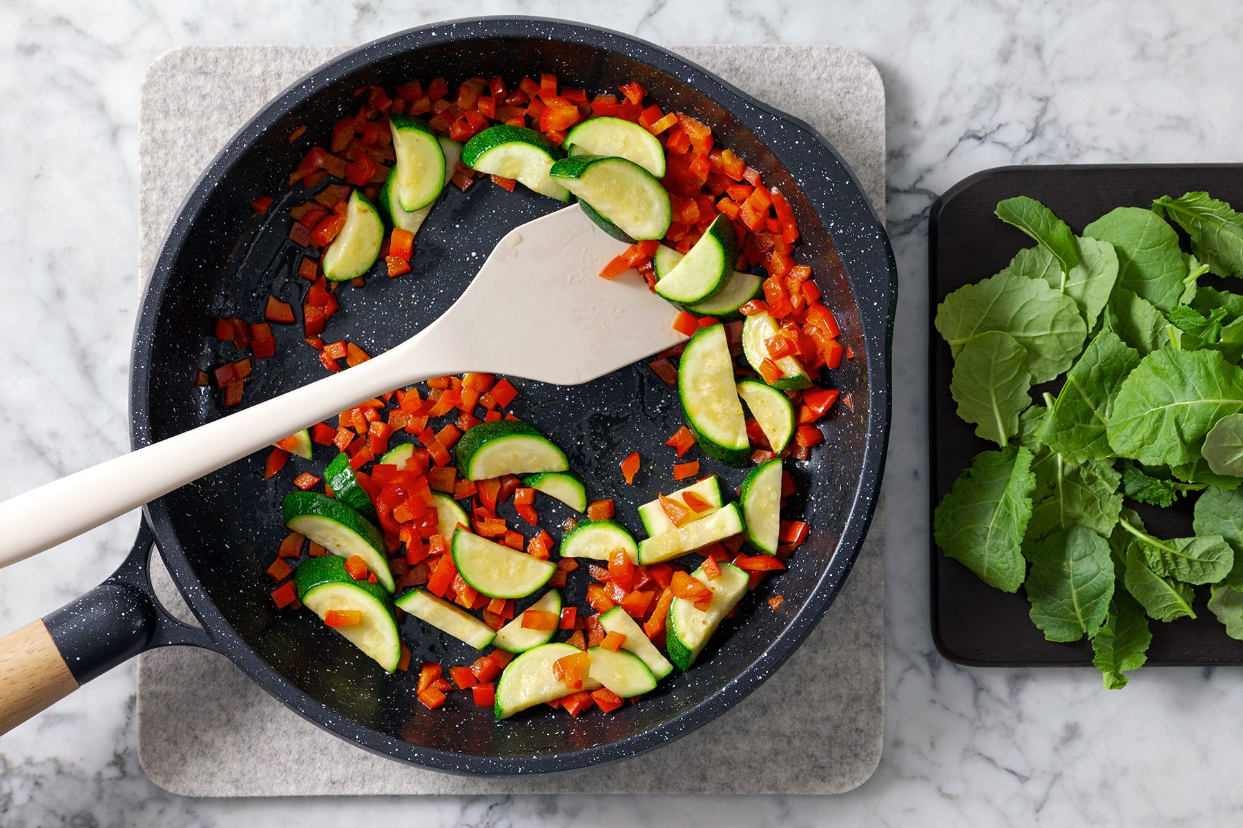 A skillet on a marble surface contains chopped zucchini and red bell peppers being stirred with a spatula. A cutting board with fresh greens is next to the skillet.