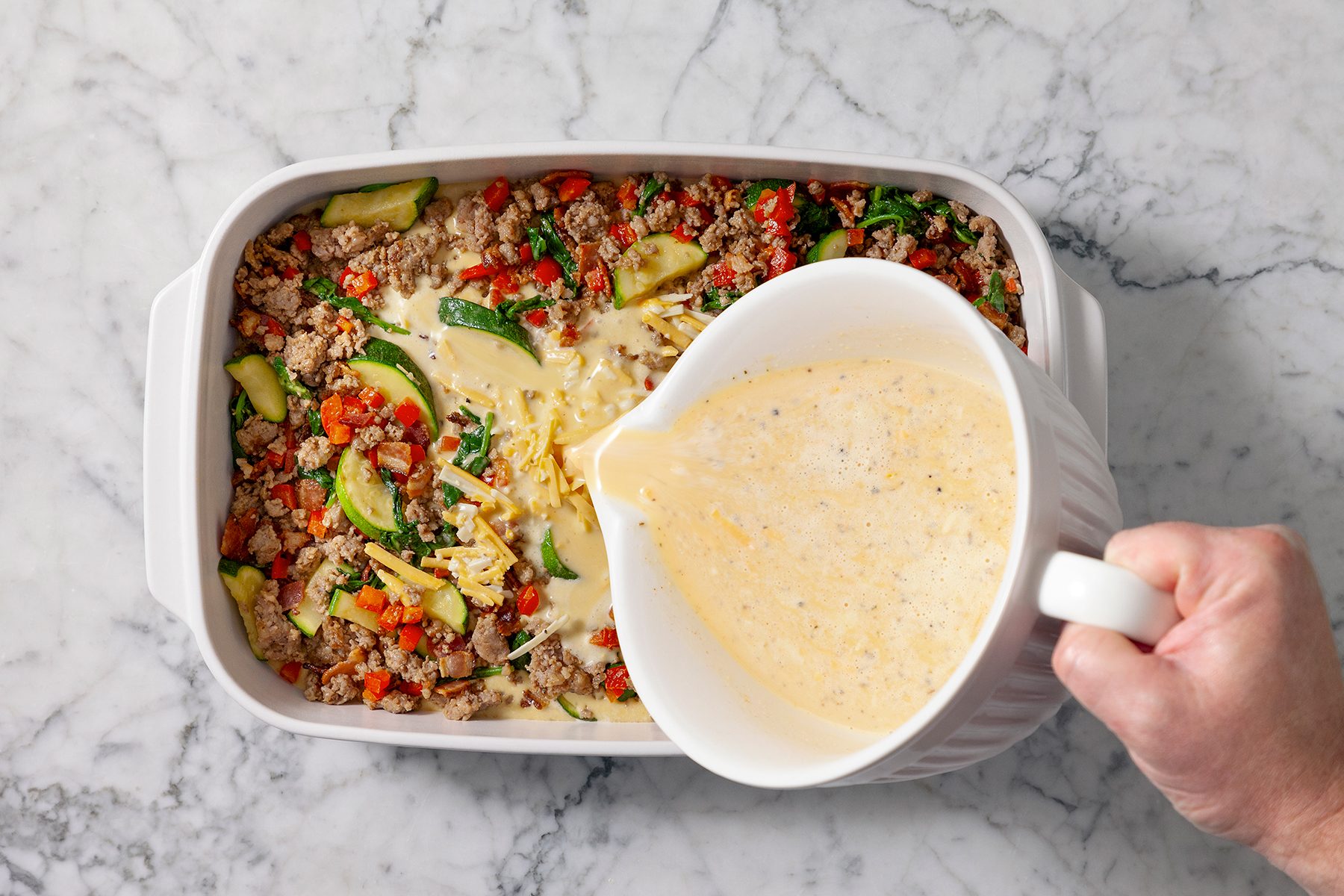 A person pours a creamy egg mixture over a casserole dish filled with ground meat, zucchini slices, chopped peppers, and spinach on a marble countertop.