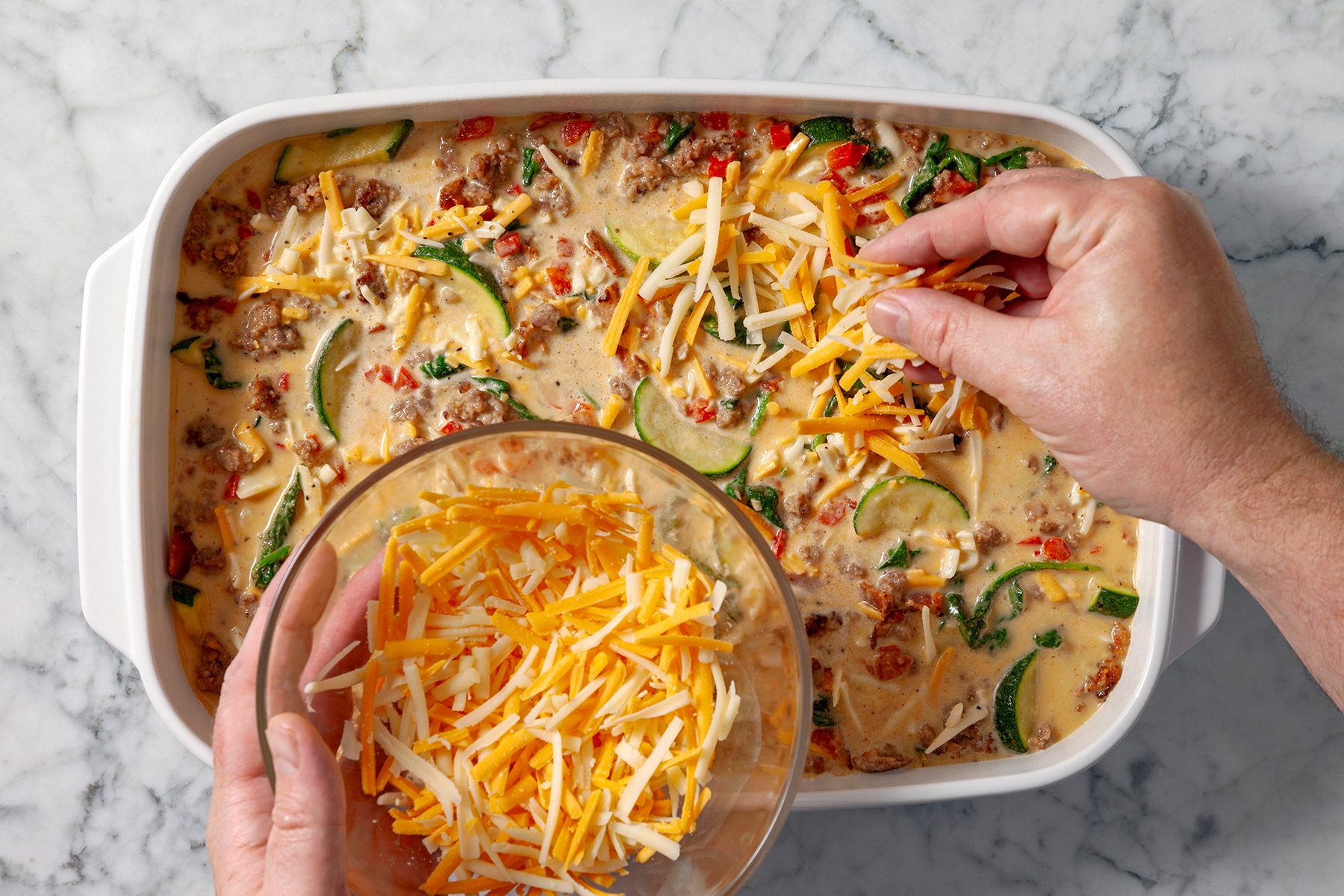 Person sprinkling shredded cheese over a casserole dish filled with a mixture of vegetables, meat, and sauce on a marble countertop.